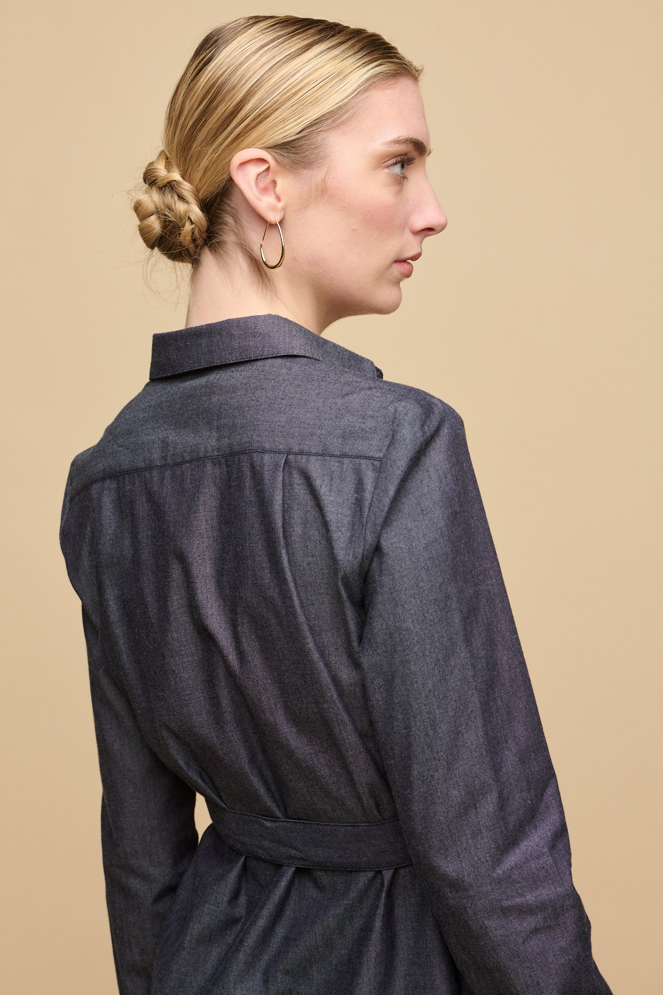 Hip up image of the back of female with blonde hair tied into a low bun wearing collared shirt dress in indigo