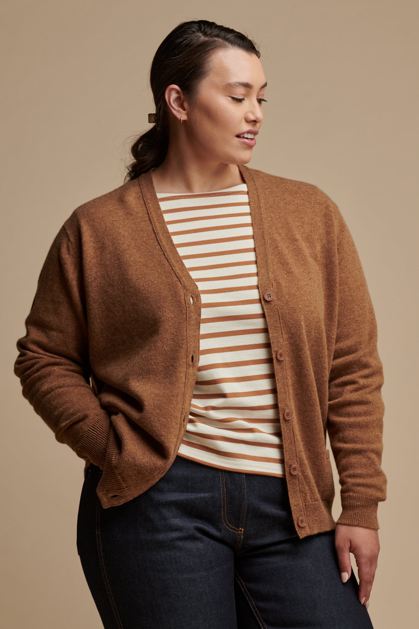 Thigh up image of female with her hand in the front jeans pocket wearing lambswool v neck cardigan in dark camel unbuttoned over Breton top in ecru-cinnamon, paired with indigo jeans.