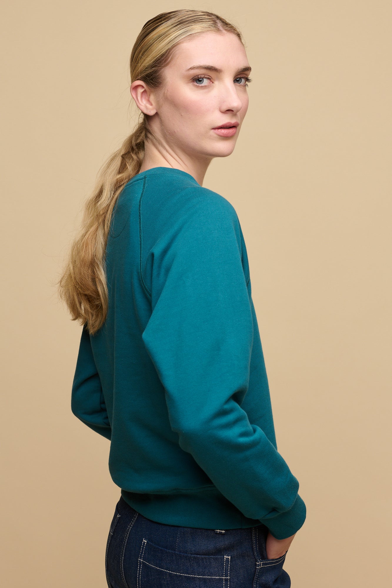The side of female with blonde hair tied in low ponytail wearing raglan sweatshirt in teal paired with work jeans in blue