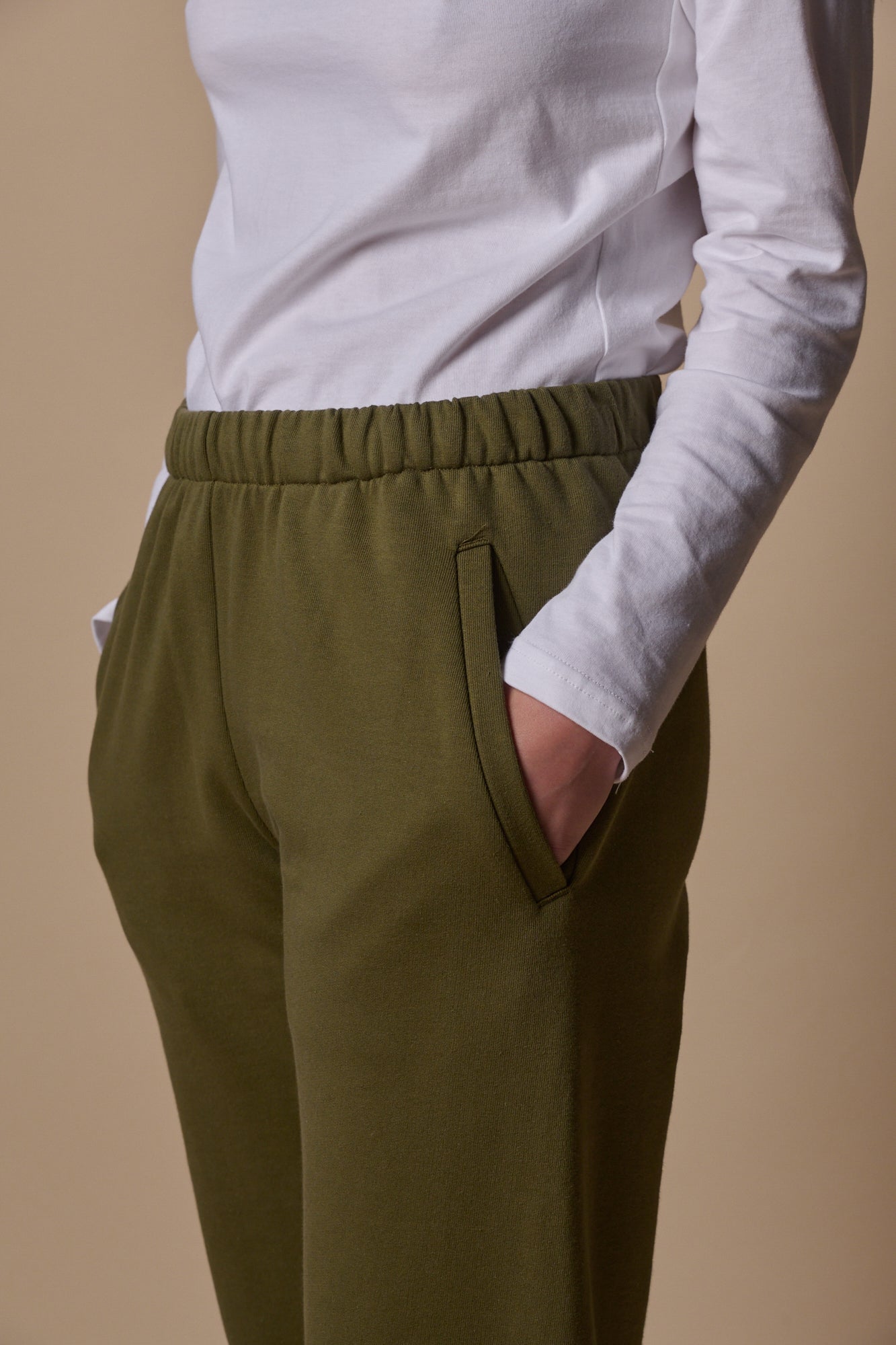 close up showing elasticated waistband of sweatpants in olive with two front pockets, model wearing long sleeve t shirt tucked in