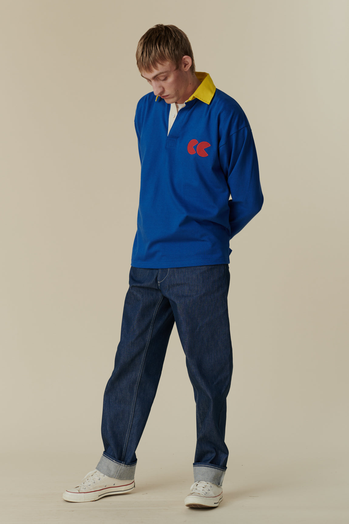 Full body front of white male wearing long sleeve blue rugby shirt with contrast yellow collar and red CC logo badge on chest, worn over plain white t shirt.