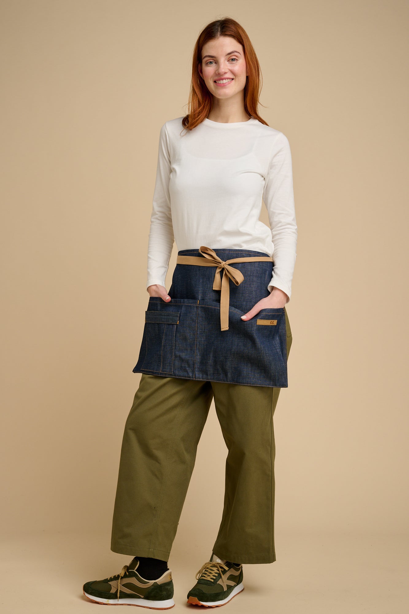 Woman wearing a white t shirt, denim apron, and green pants on a beige background.