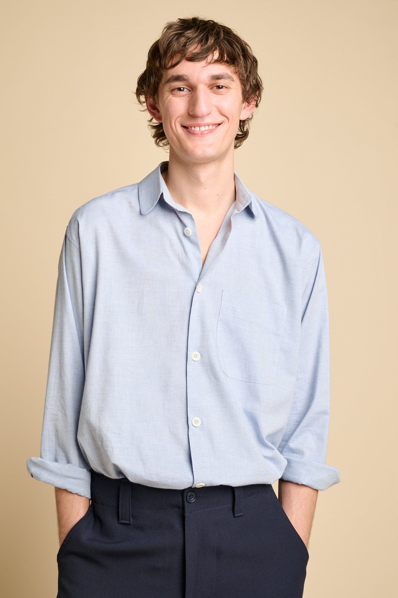 A smiley man wearing long sleeve light blue shirt tucked into navy trousers.