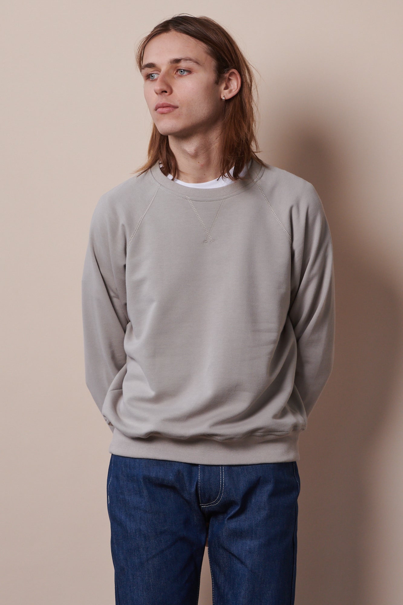 Thigh up image of white male with shoulder length brunet hair wearing raglan sweatshirt in stone paired with straight cut jeans in blue
