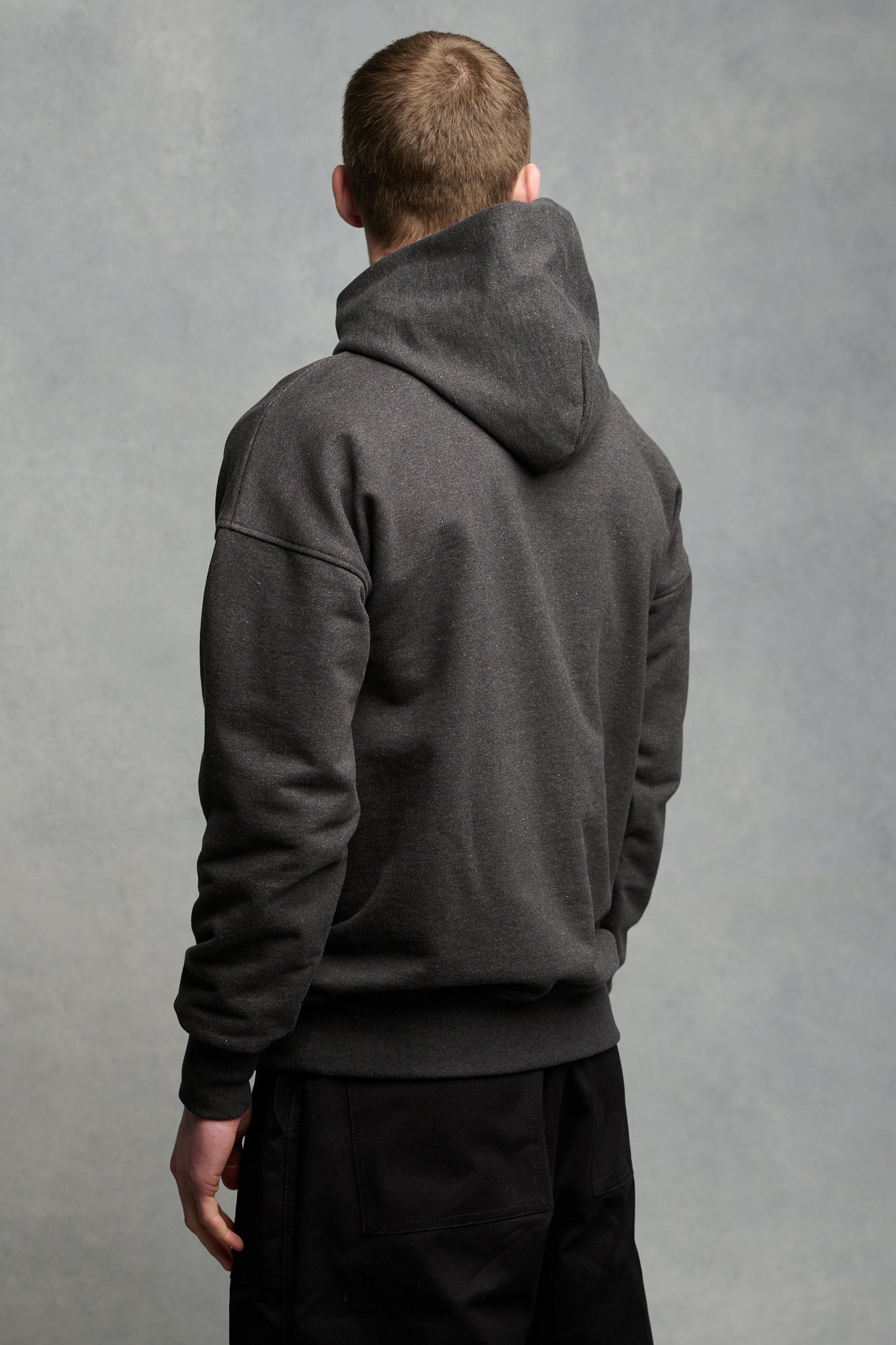 Thigh up image of the back of brunet male wearing heritage hooded sweatshirt in charcoal paired with cameraman pants in black