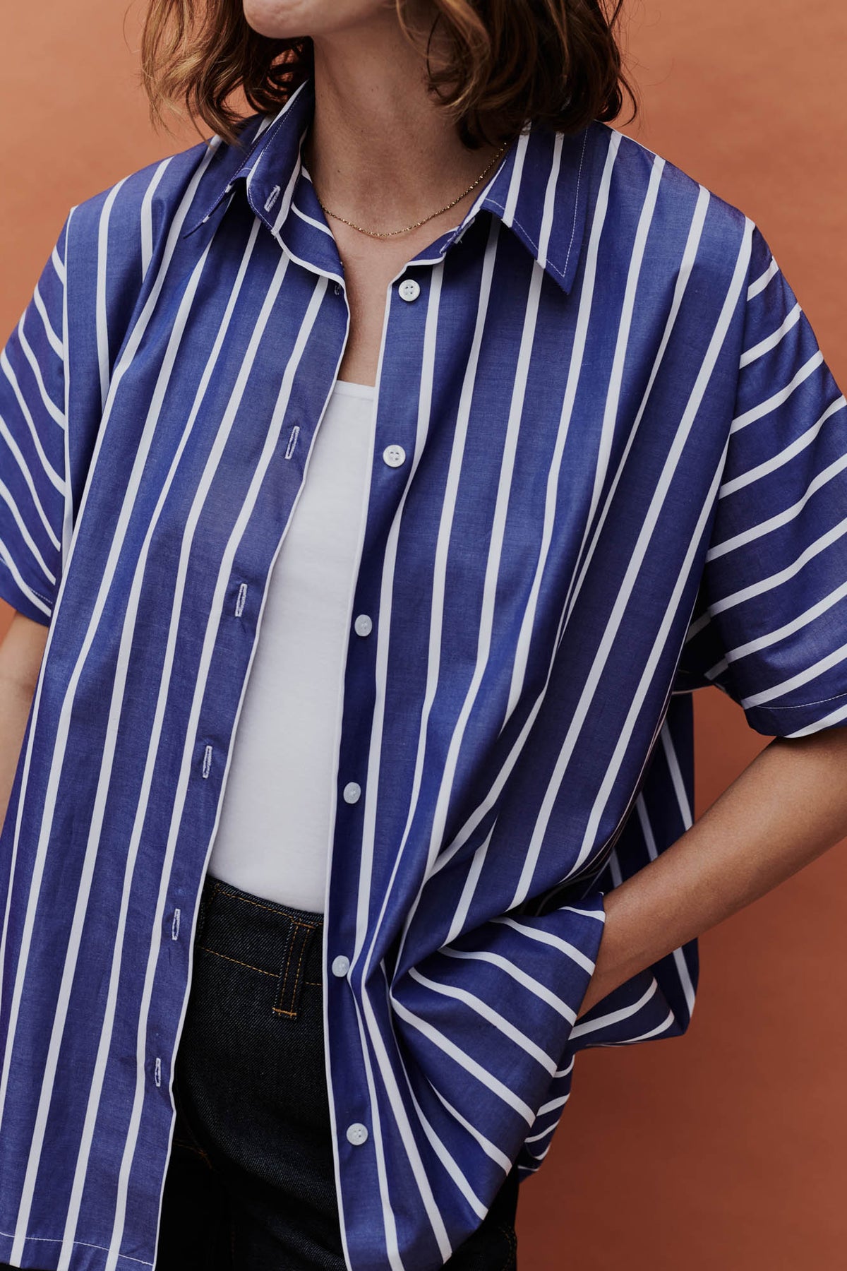 female wearing unbuttoned Ava short sleeve shirt with wide leg jeans with a white camisole in front of a coral background