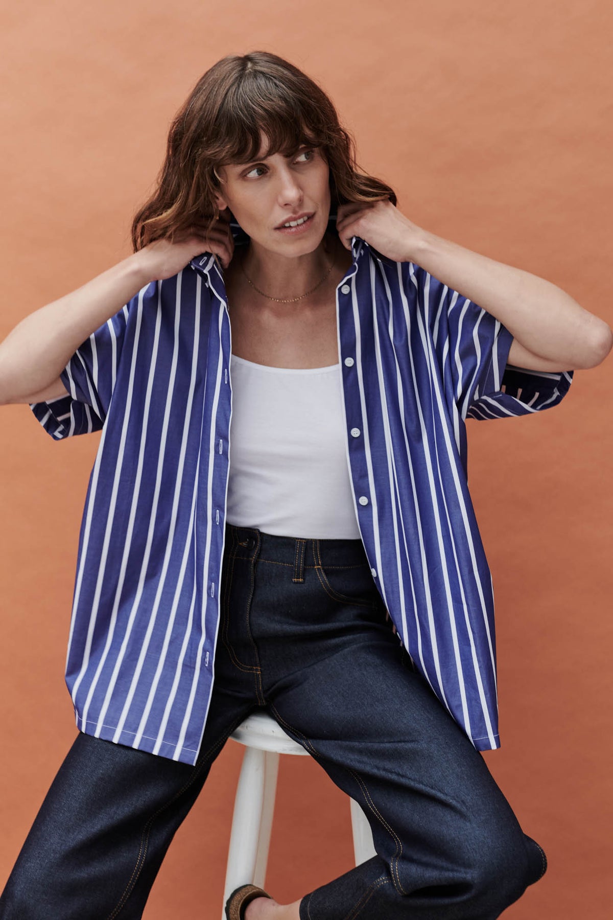 female wearing unbuttoned Ava short sleeve shirt with wide leg jeans with a white camisole in front of a coral background