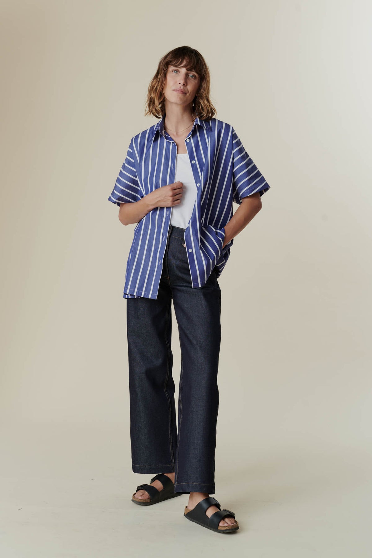 female wearing unbuttoned Ava short sleeve shirt with wide leg jeans with a white camisole in front of a beige background