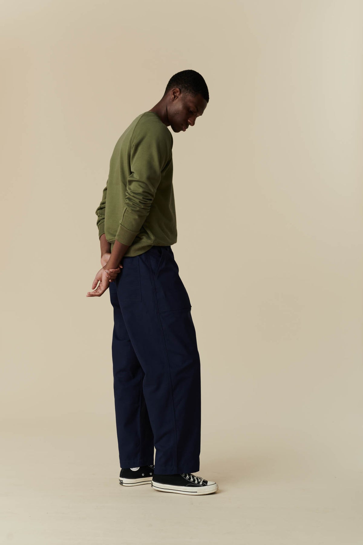 Side profile view of a man wearing olive raglan sweatshirt with navy tapered fit cameraman pant with black 70s Converse.
