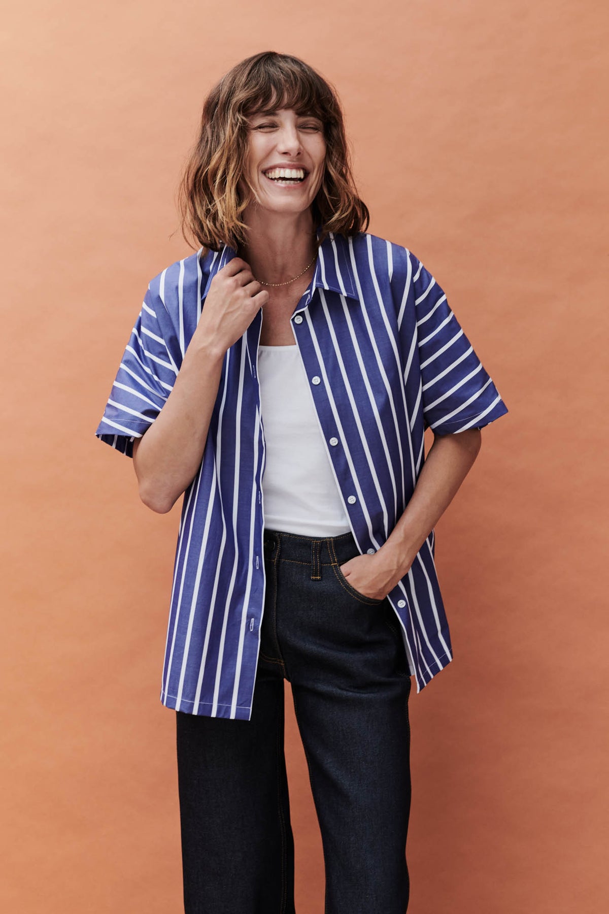 female wearing unbuttoned Ava short sleeve shirt with wide leg jeans with a white camisole in front of a coral background