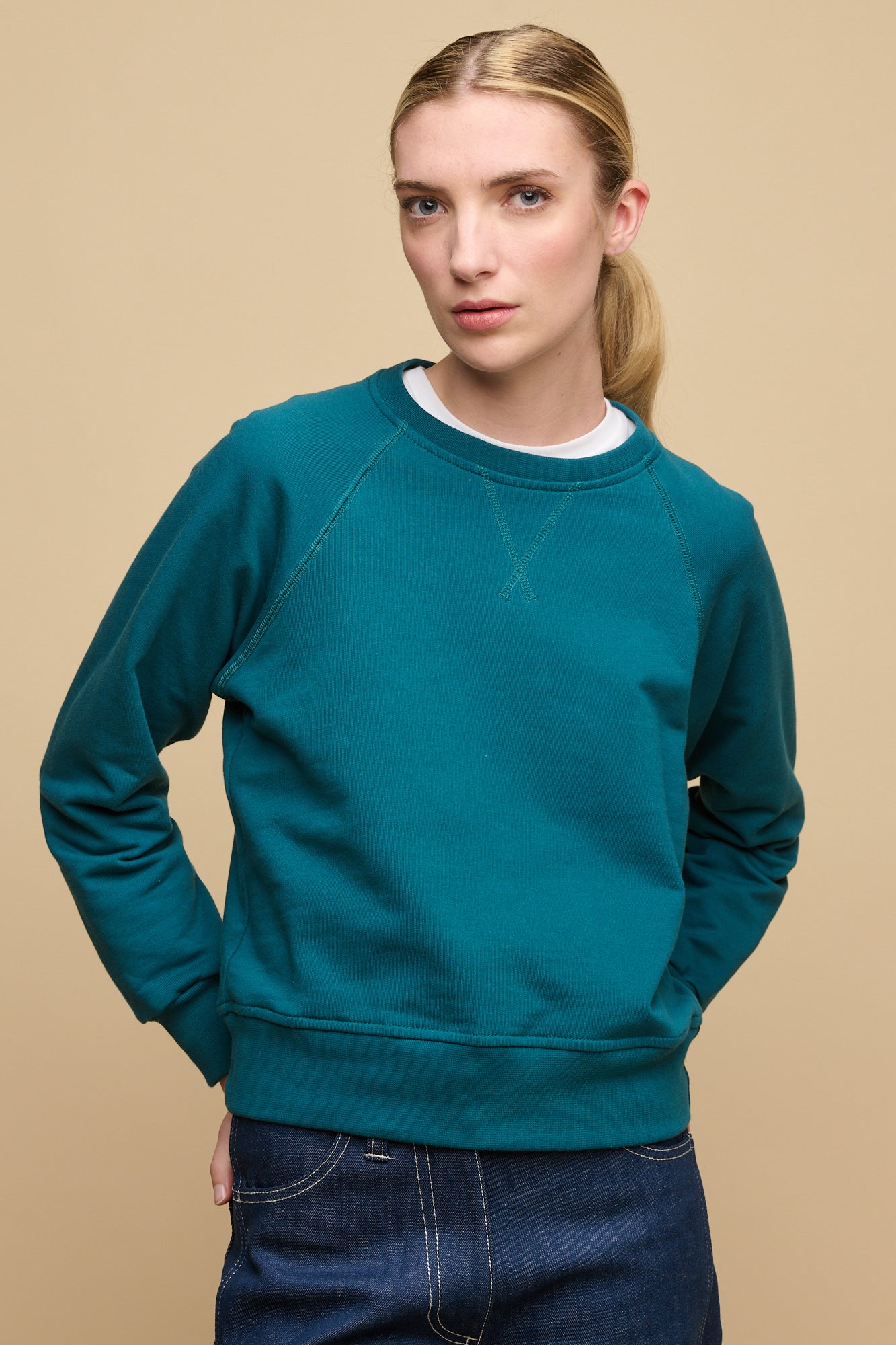 Thigh up front of female wearing Raglan sweatshirt in teal layered over crew neck t shirt in white worn with blue work jean