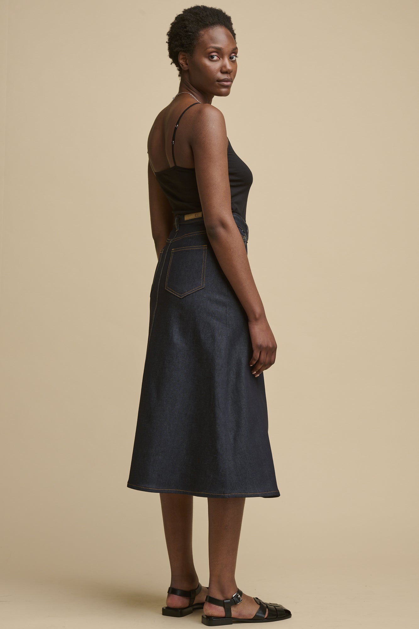 Full body image of the back of female looking over her shoulder to the camera wearing Bobbie a line denim skirt in indigo with two back pockets and C logo patch on the waistband