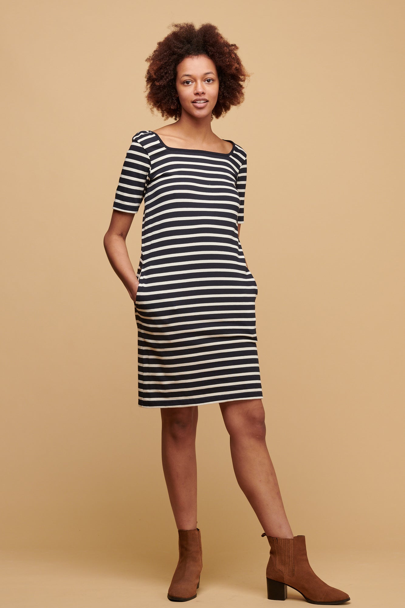 Female of colour with afro tilting her head to the side wearing navy ecru stripe Breton dress with navy binding round the square neck.