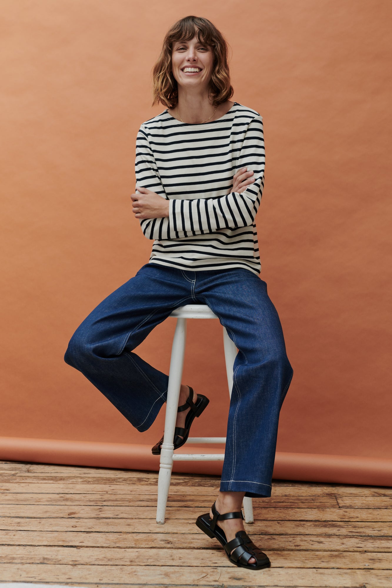 Brunette female sat on stool wearing Breton in ecru navy stripe paired with work jeans in blue