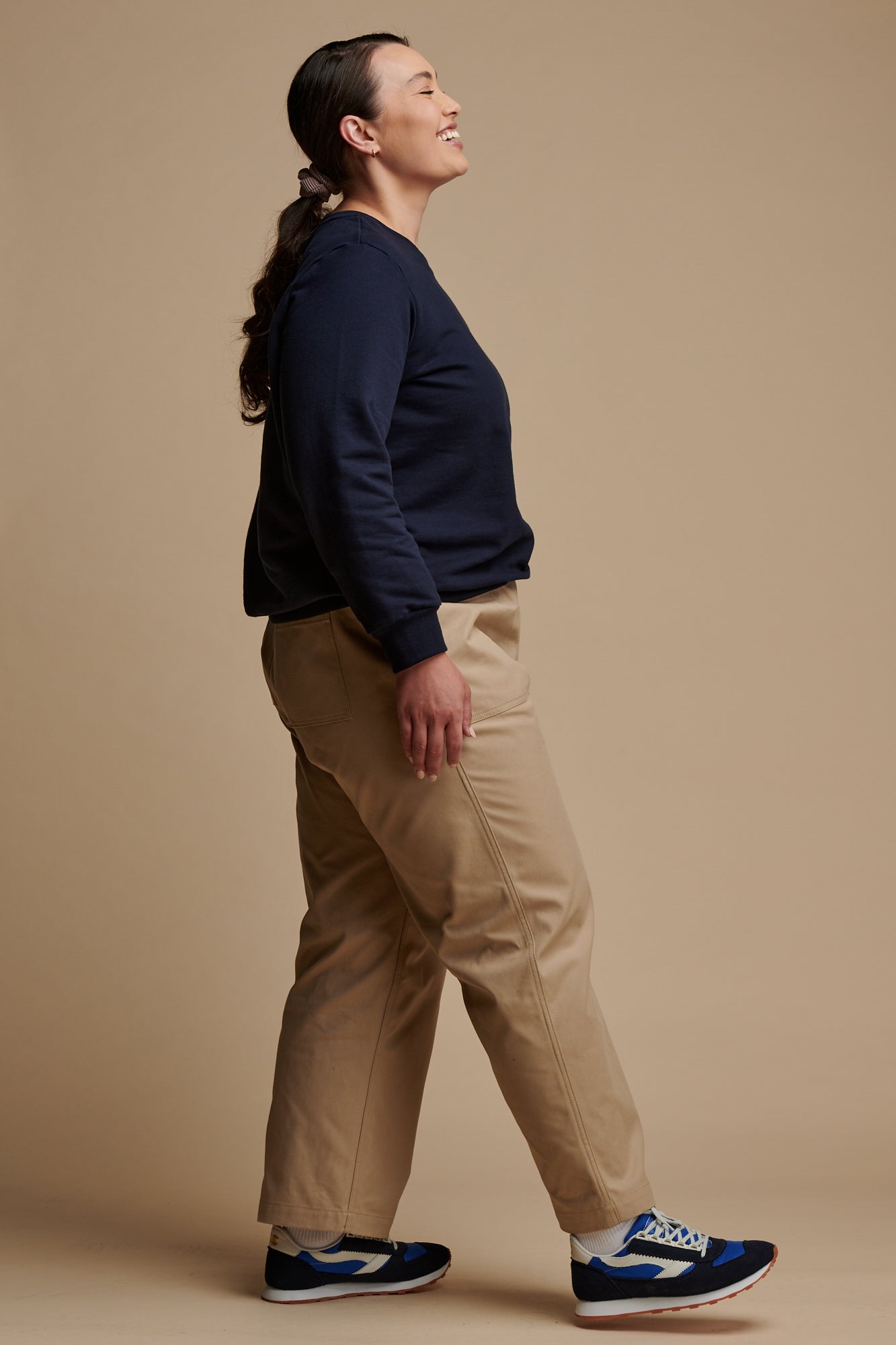The side of smiley female wearing camerawoman drawstring tapered cotton canvas trousers in putty paired with navy sweatshirt and Community Clothing X Walsh trainers.