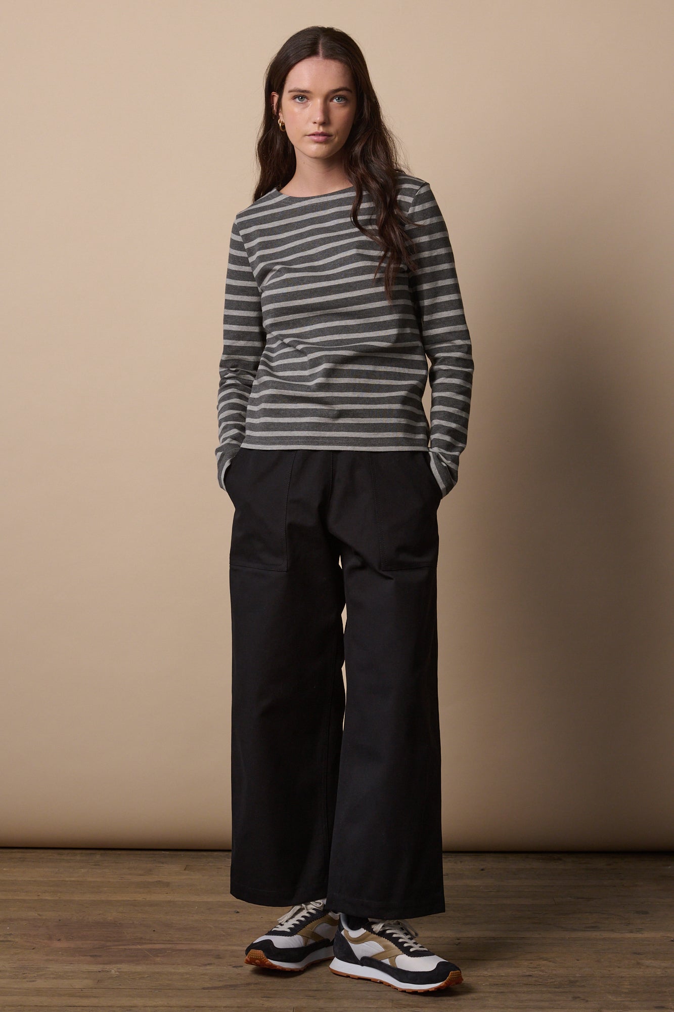 Full body image of the front of female wearing camerawoman cotton trousers in black with hands in the two front pockets, paired with long sleeve breton
