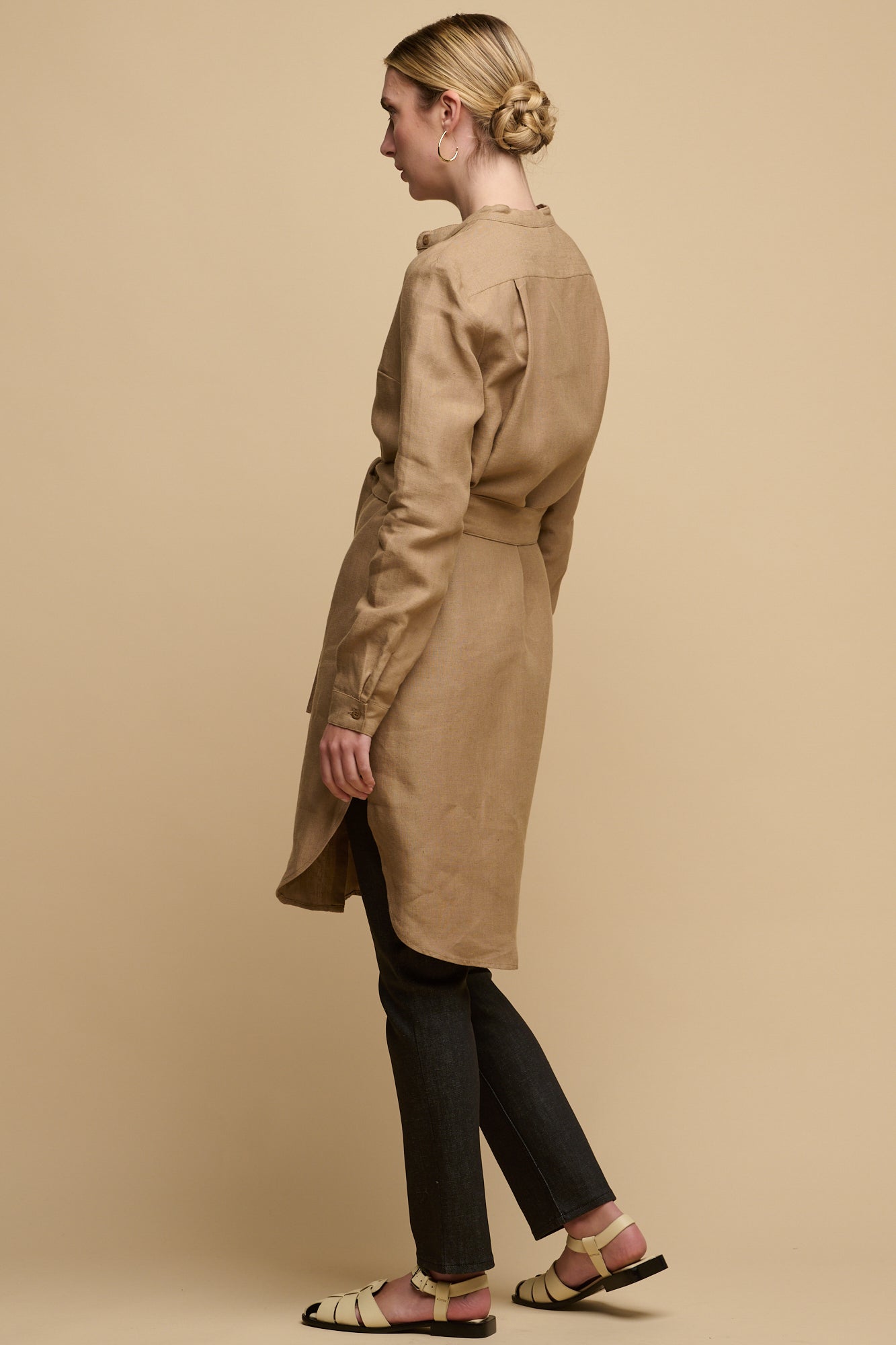 Full body image of the back of female wearing collarless shirt dress in truffle linen with waist belt tie. Dress worn over straight leg jean in black paired with sandals for a day time look.