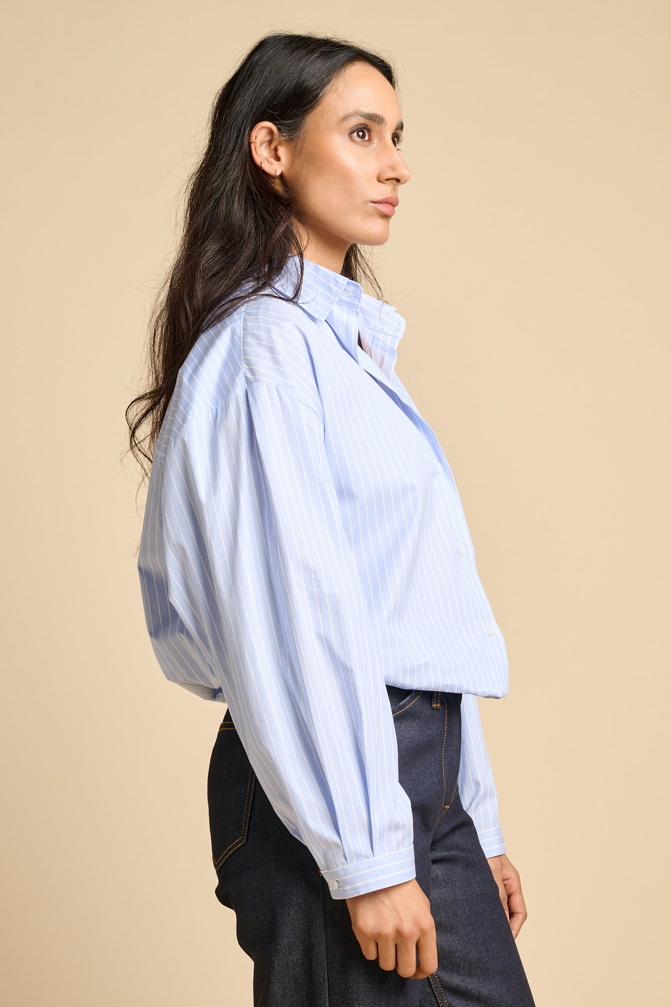 Side profile of a woman wearing long sleeve blue and white stripe Esme shirt tucked into indigo jeans.