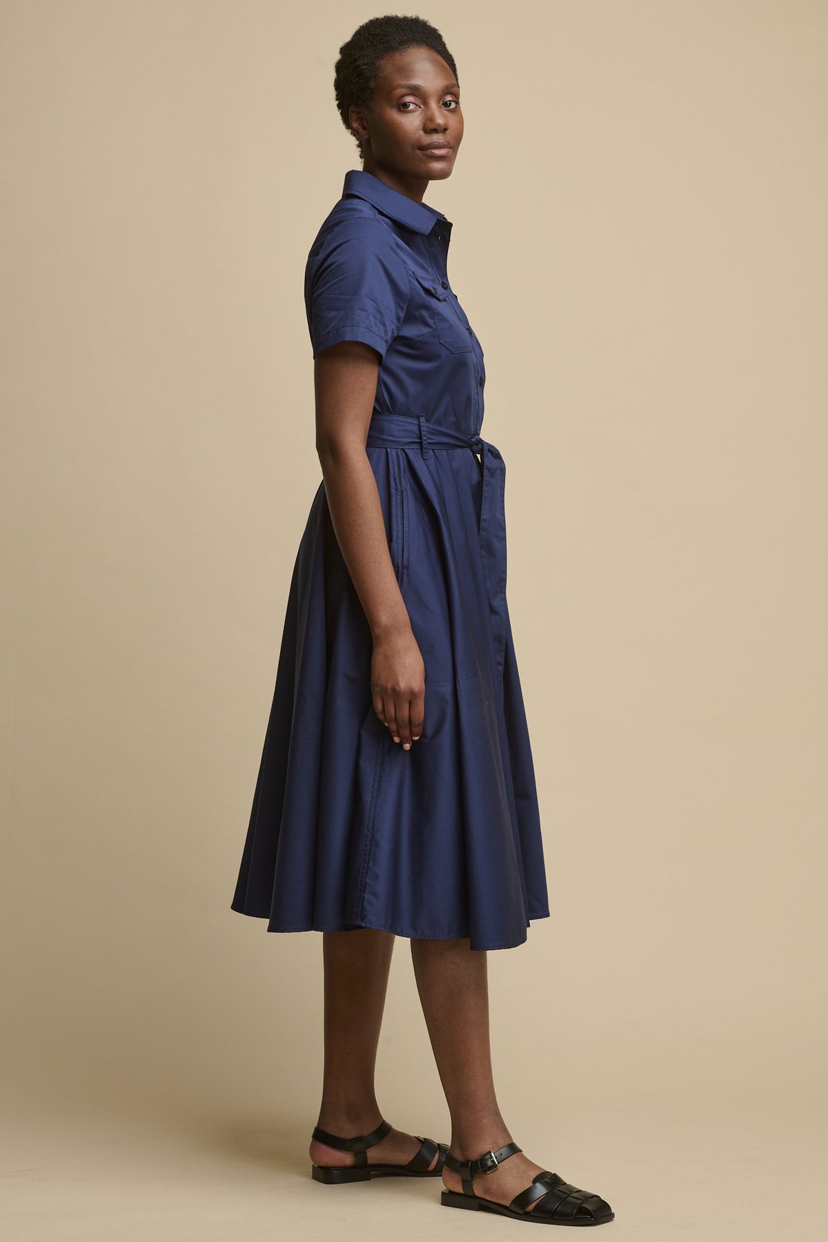 Full body image of the side of female wearing full skirted phoebe trench dress in navy paired with black sandals 