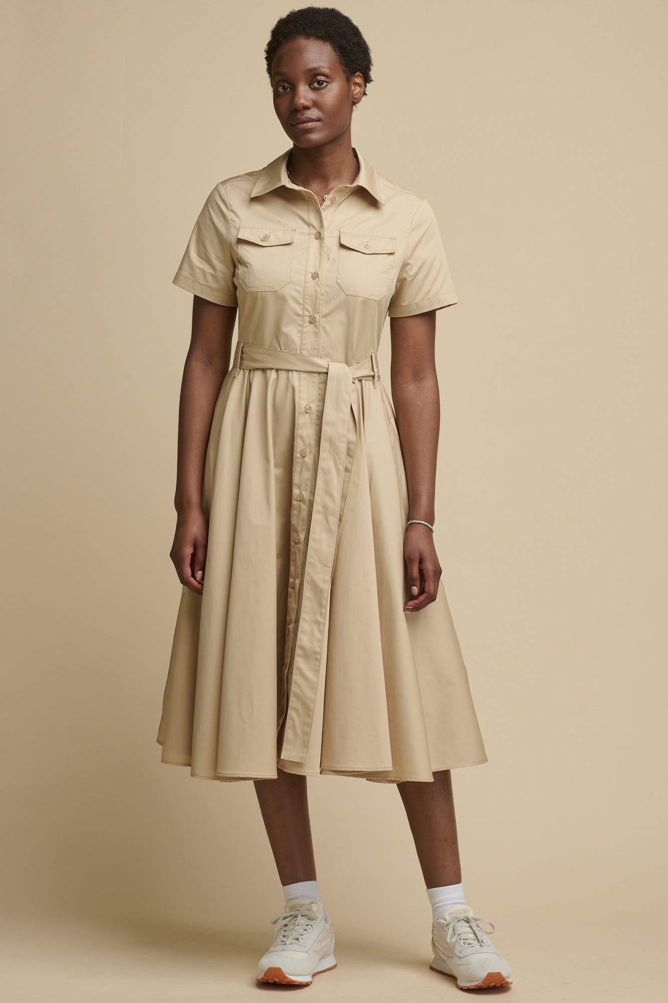 Full body image of the front of female wearing full skirted phoebe trench dress in stone paired with trainers