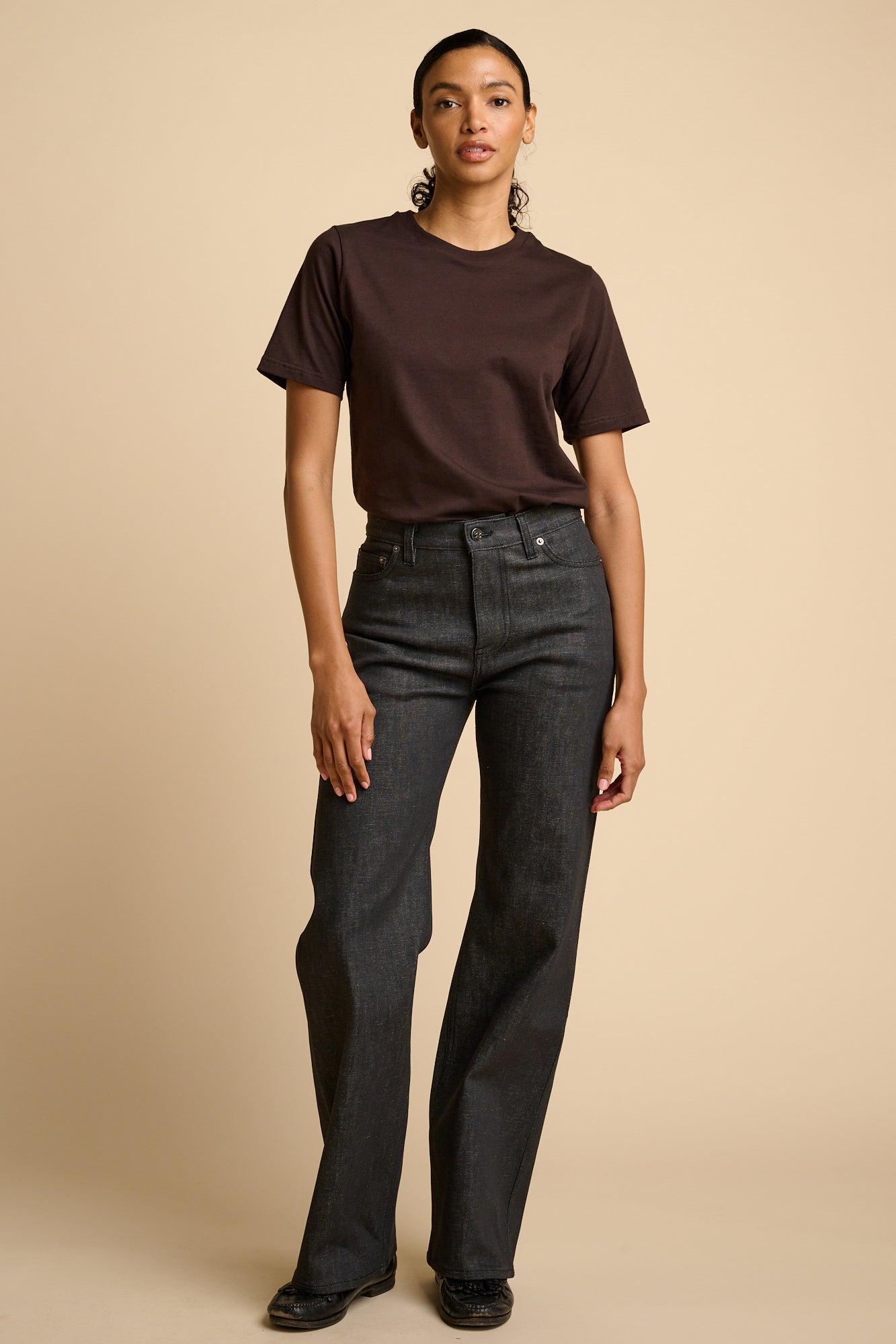 Woman wearing a brown t-shirt and black jeans on a beige background.
