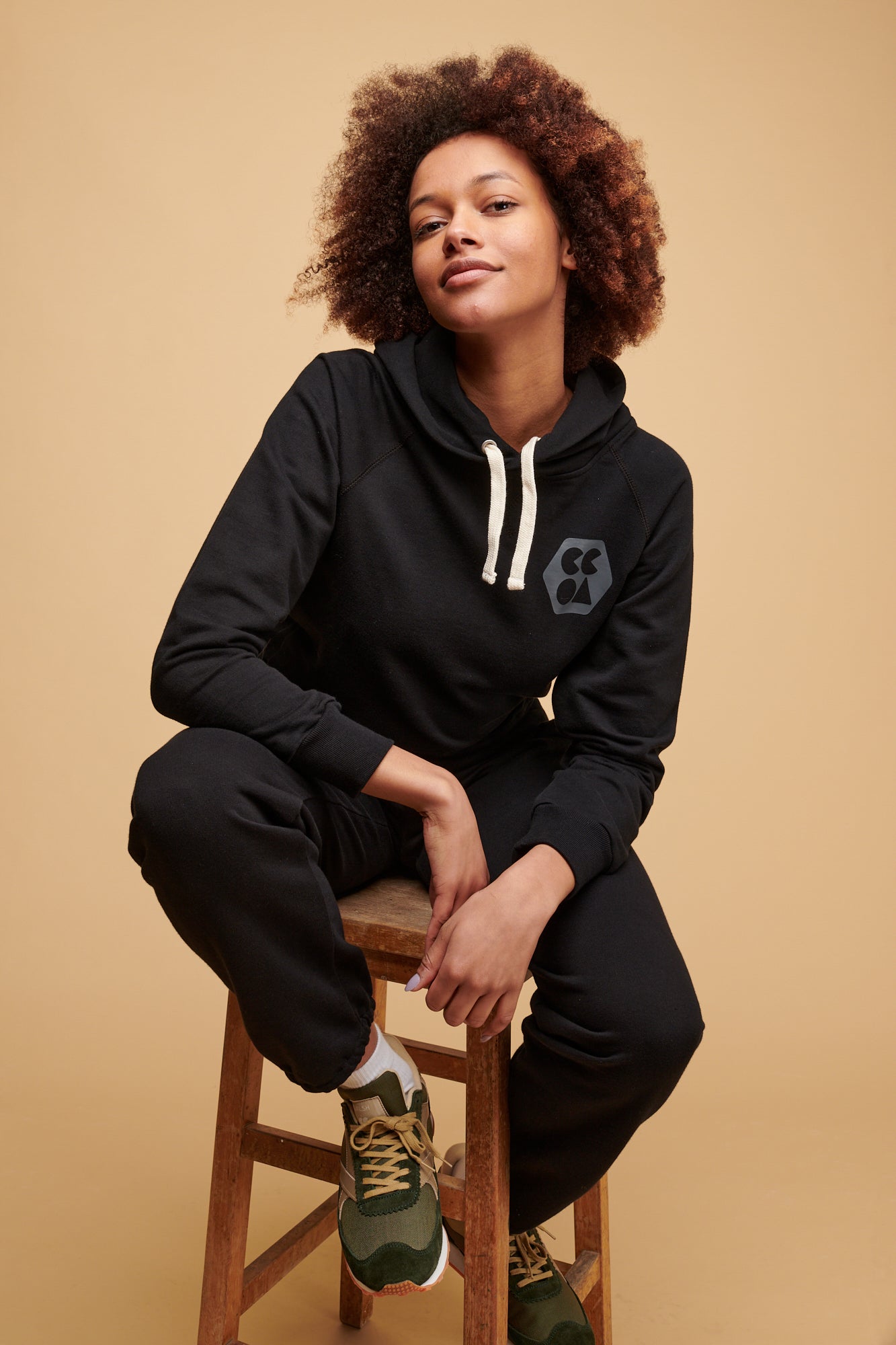 Female of colour sat on stool wearing plastic free hooded sweatshirt in black with ecru drawcord through the hood and CCOA logo print on chest paired with 100% cotton sweatpants in black