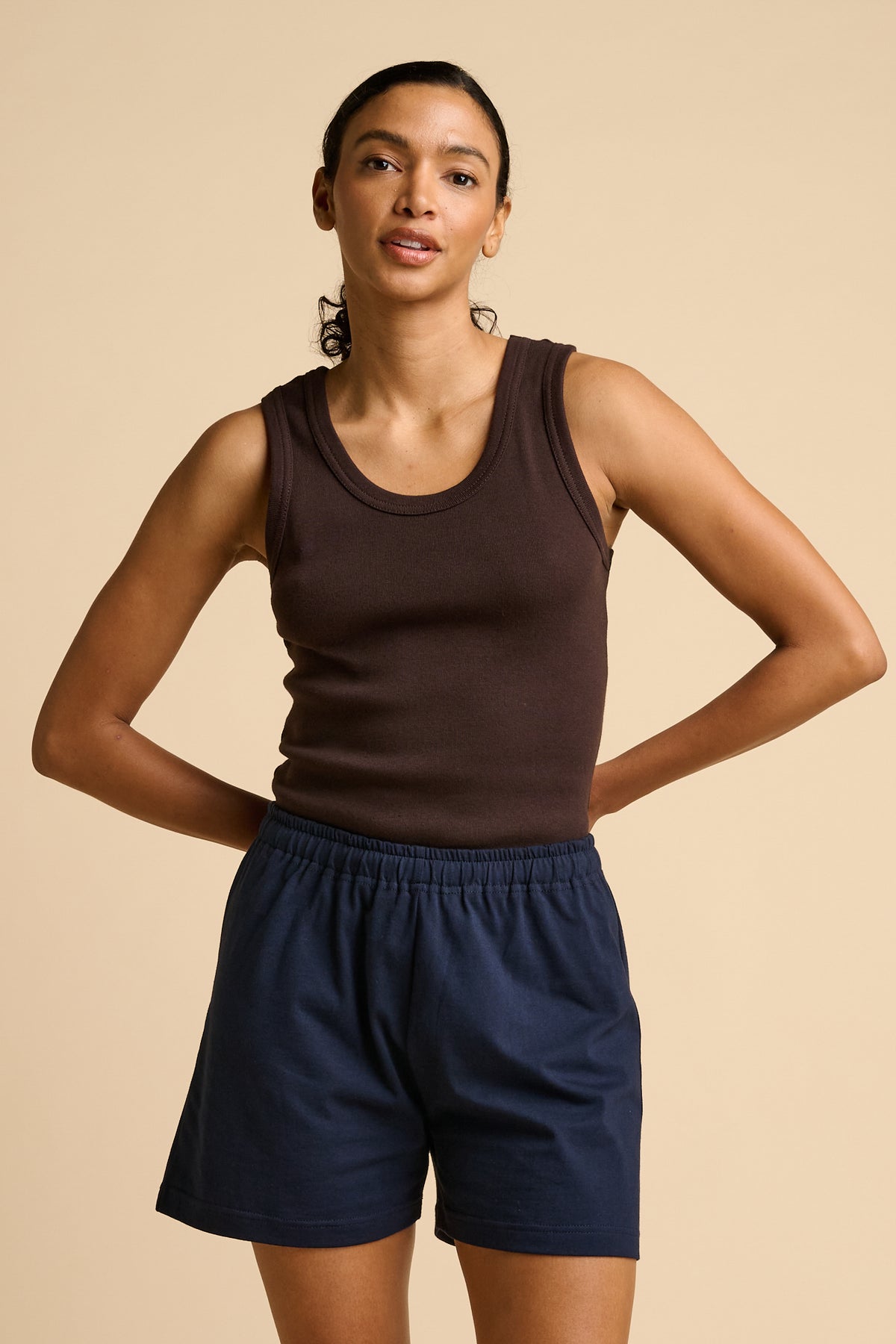 Woman wearing a brown tank top and navy shorts on a beige background.