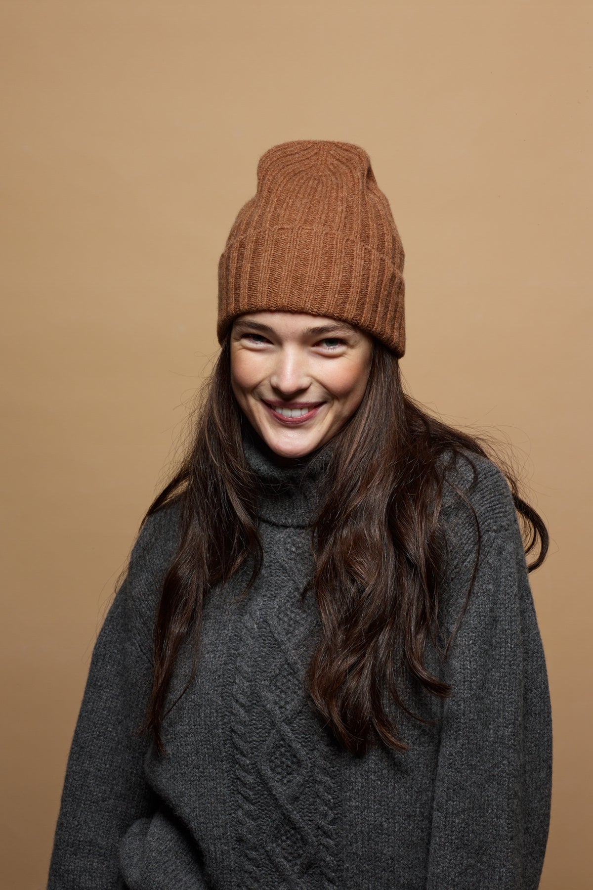 Smiley, brunette female wearing unisex lambswool beanie hat in dark camel