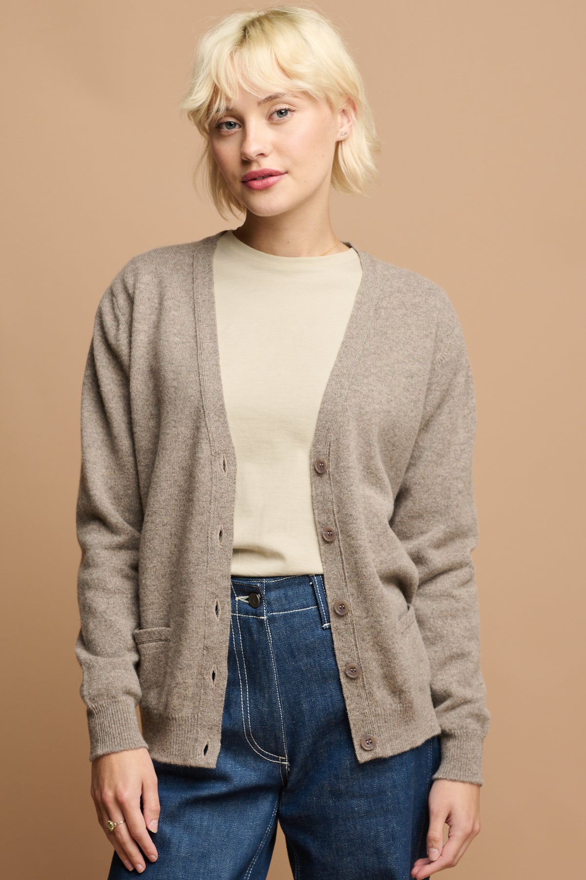 Thigh up image of female wearing v neck lambswool cardigan in truffle unbuttoned over crew neck t shirt in stone tucked into work jeans in blue