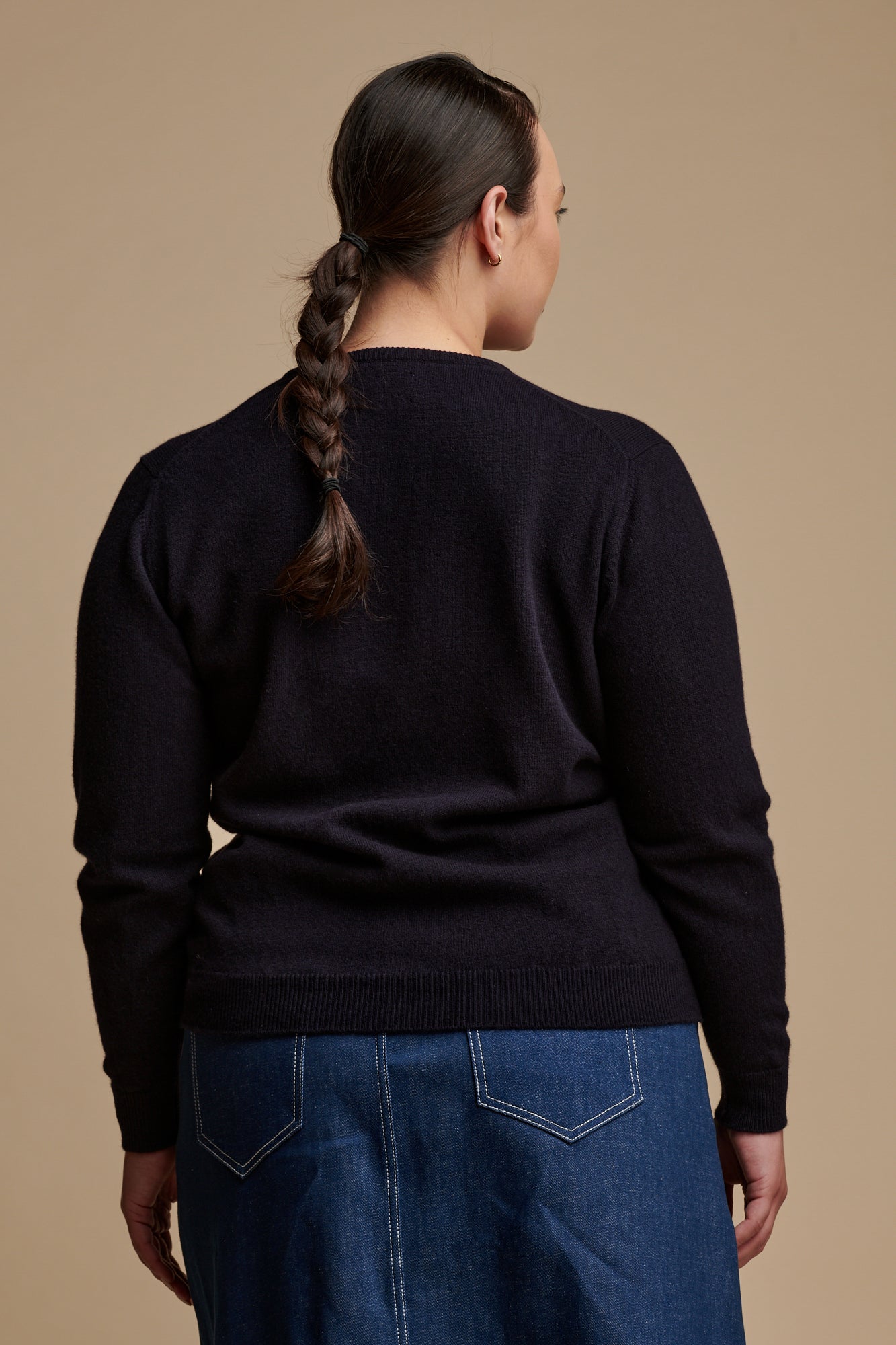 Thigh up of the back of female with dark hair, tied back into a low ponytail plait, wearing lambswool crew neck jumper in navy paired with Bobbie a line denim midi skirt in blue.