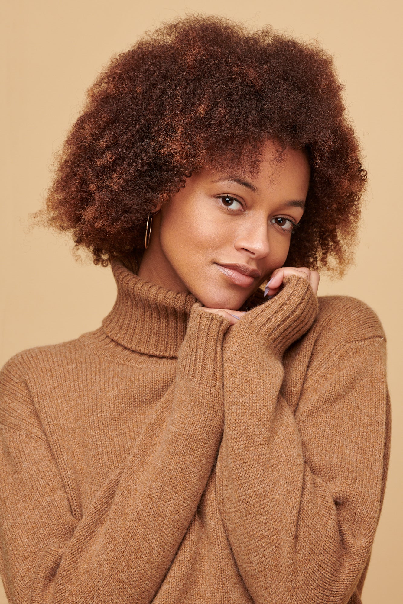 Portrait of female of colour with an afro wearing lambswool roll neck in dark camel holding her hands up under her chin