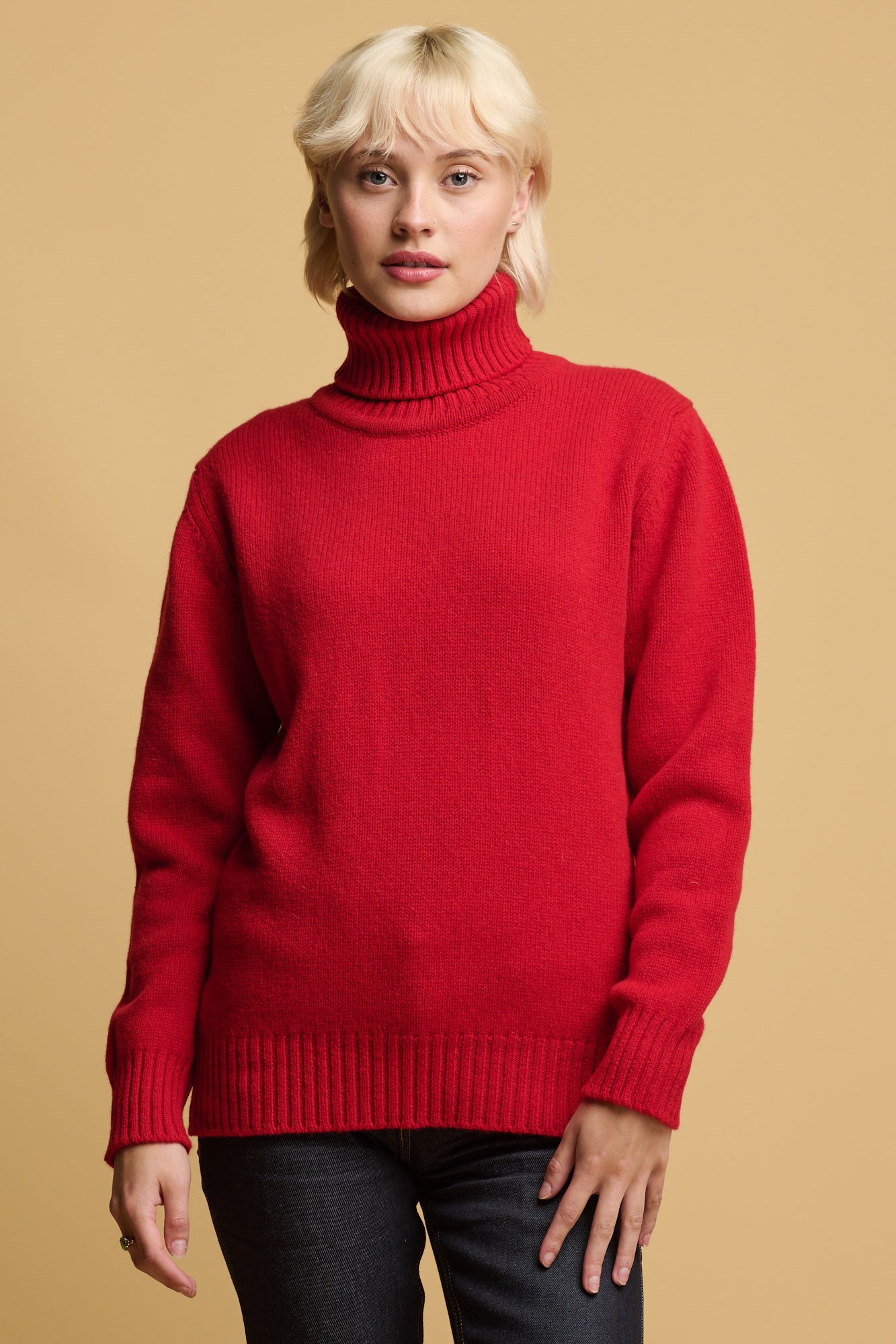 Thigh up of blonde female wearing lambswool roll neck in red with ribbed neck, cuff and hem paired with indigo jeans