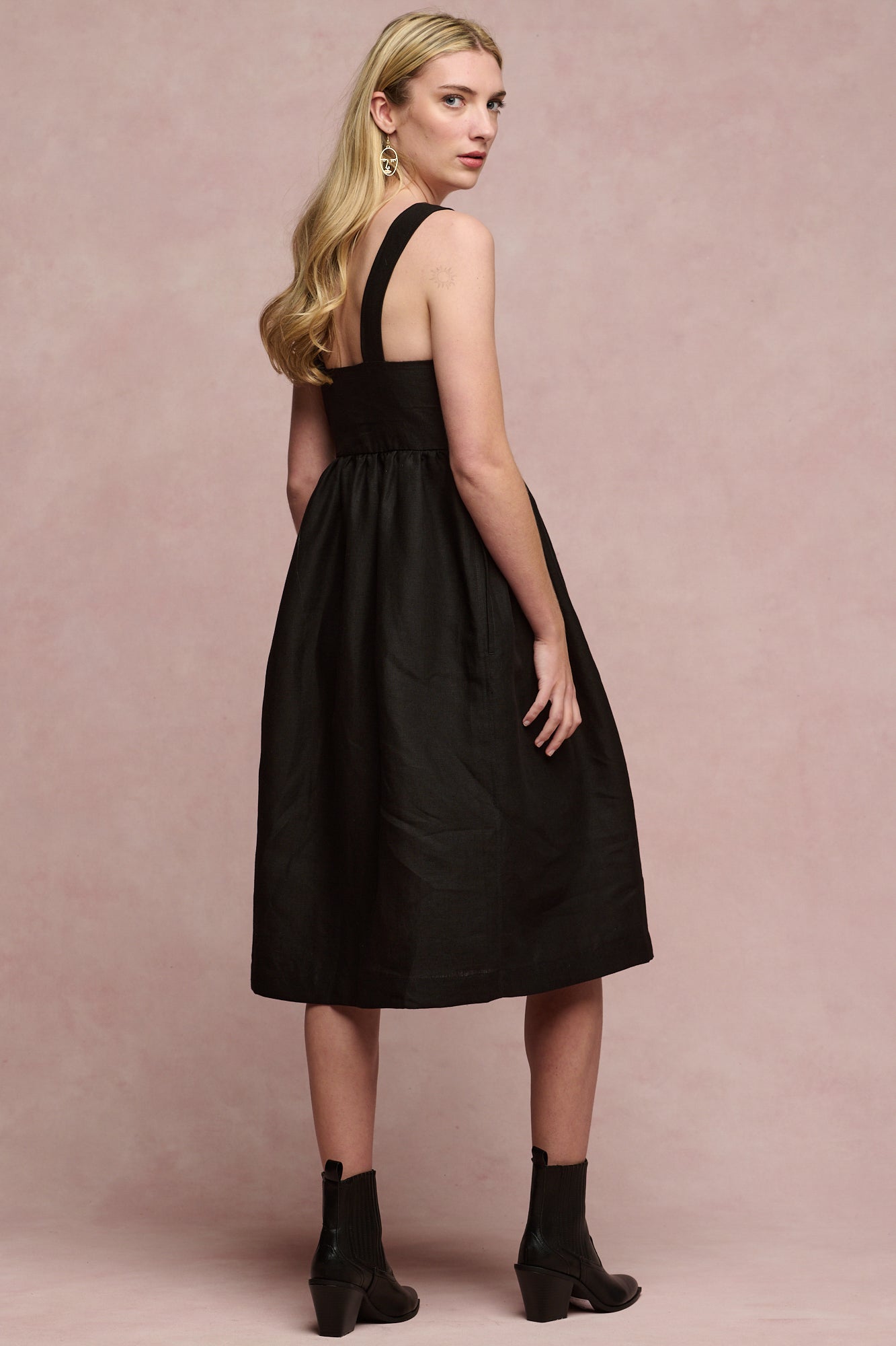 Full body image of fair skinned, blonde female wearing linen sun dress in black paired with black heeled boots
