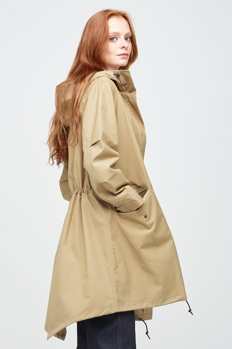 Side shot of a young, white female model with ginger hair wearing long stone coloured parka. The parka is styled with indigo jeans and a white trainer.