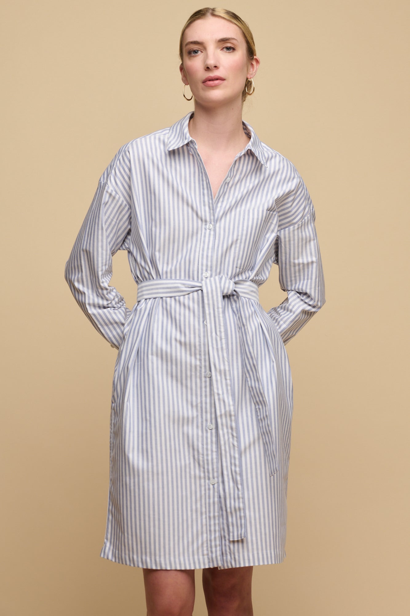 Thigh up image of white female with blonde hair tied back wearing belted midi cotton shirt dress in white blue stripes