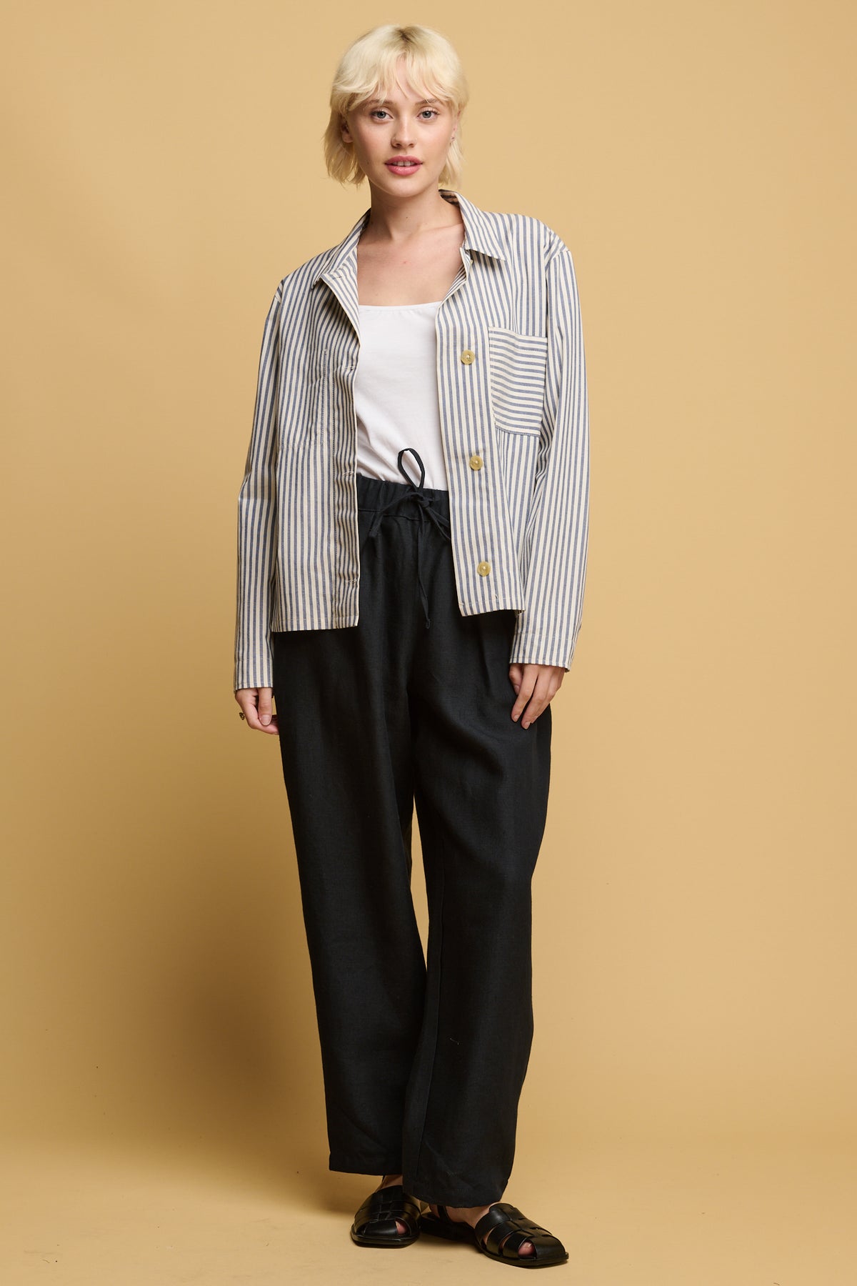front of blonde female wearing community clothing lorna  elasticated waist linen rousers in navy paired with white camisole tucked in with quarry bank blue stripe overshirt with black sandals.