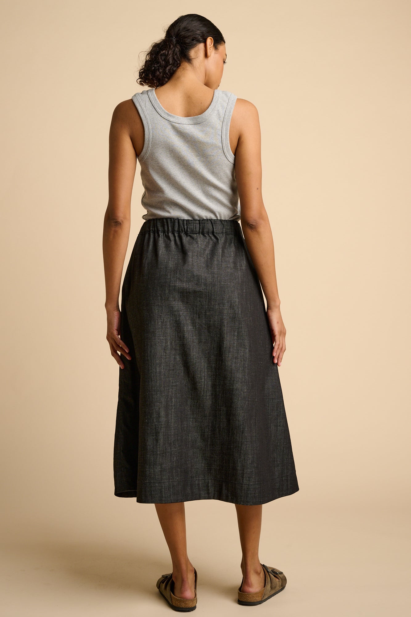 Woman wearing a grey tank top and black skirt on a beige background.