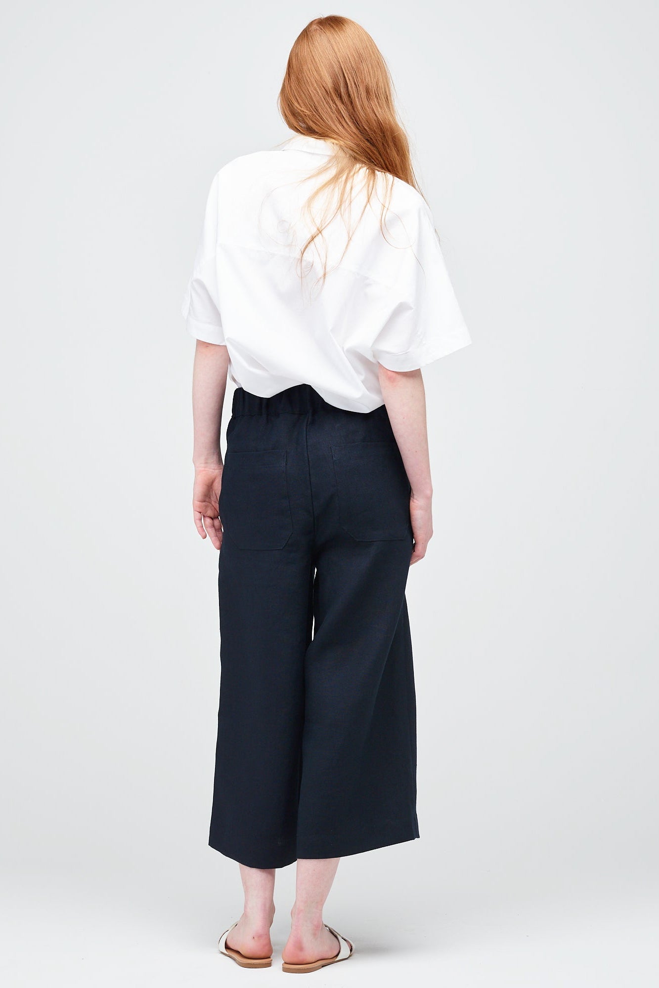 Rear shot of a young white women with ginger hair wearing short pj trouser in navy linen, styled with an oversized white shirt and sandal.