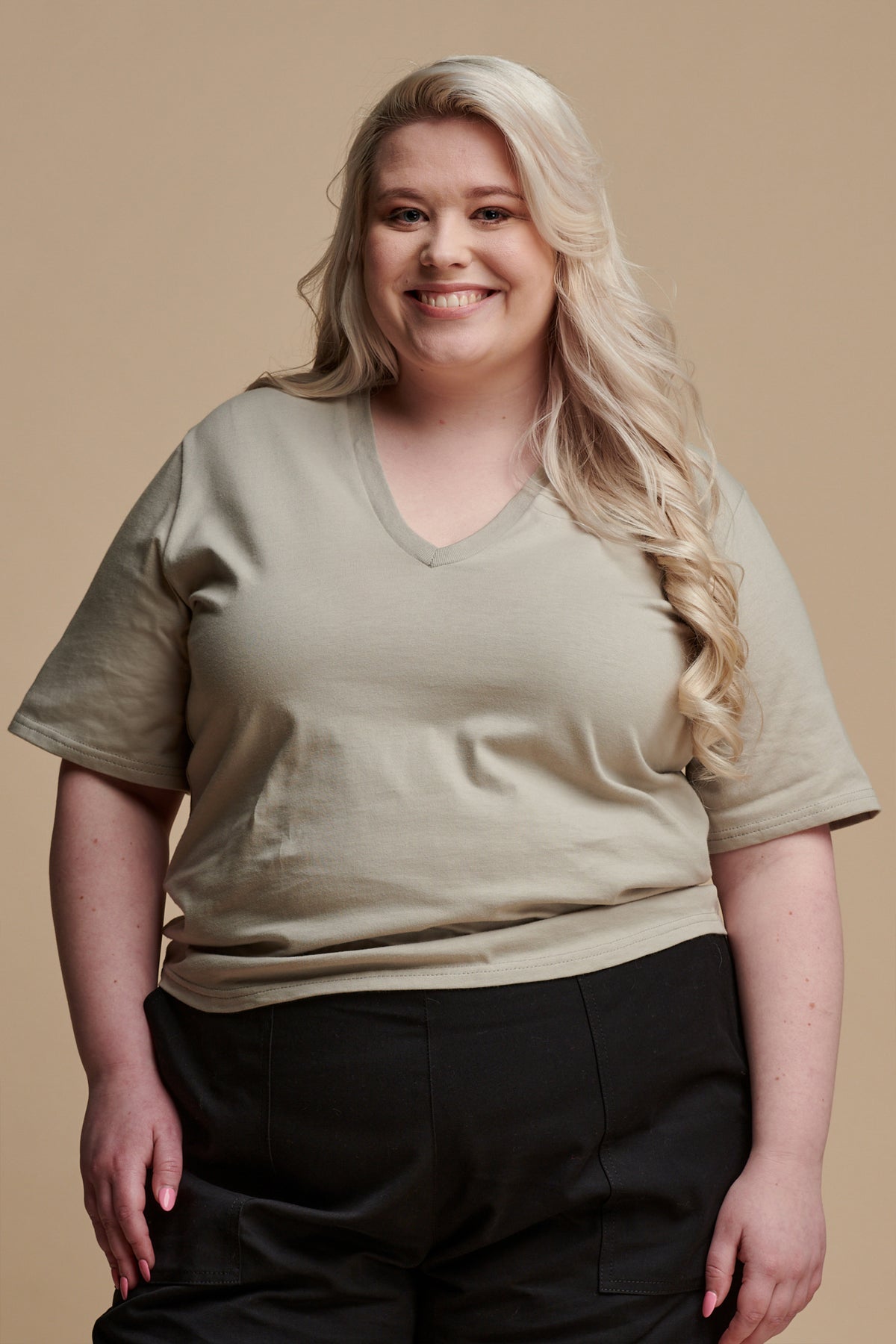 Thigh up of the front of smiley female with long, curled blonde hair wearing short sleeve v neck t shirt in stone paired with camerawoman pants in black.