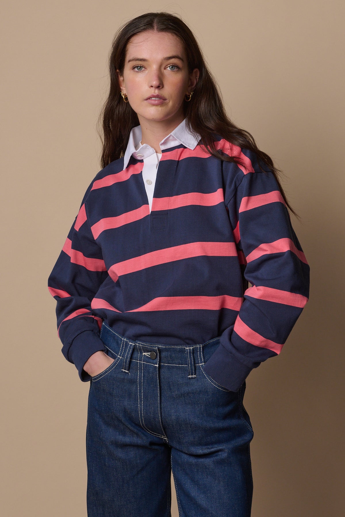 Thigh up image of white female wearing unisex striped rugby shirt in navy pink tucked into jeans