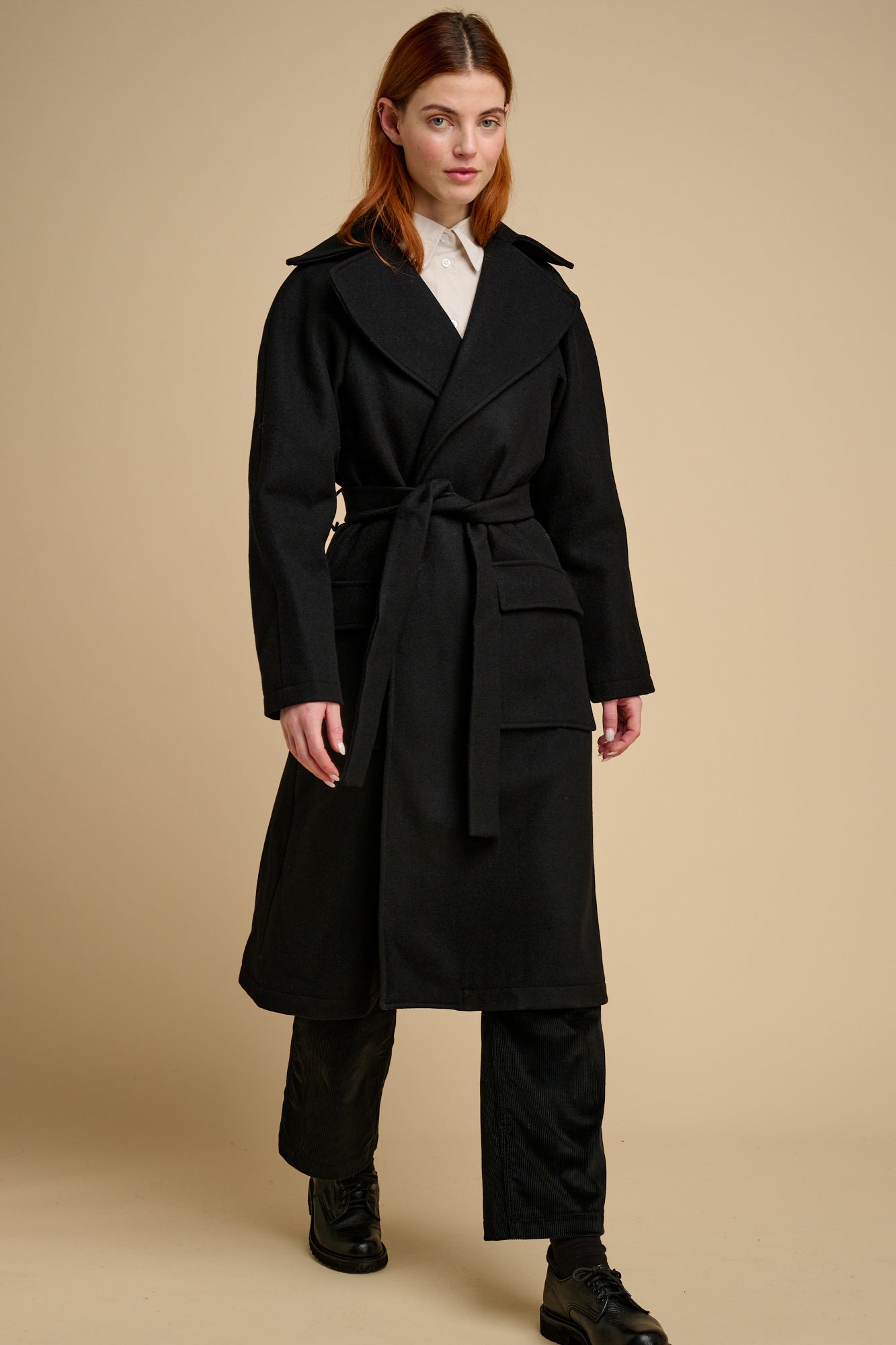 Woman wearing black wool coat with tie belt on beige background.