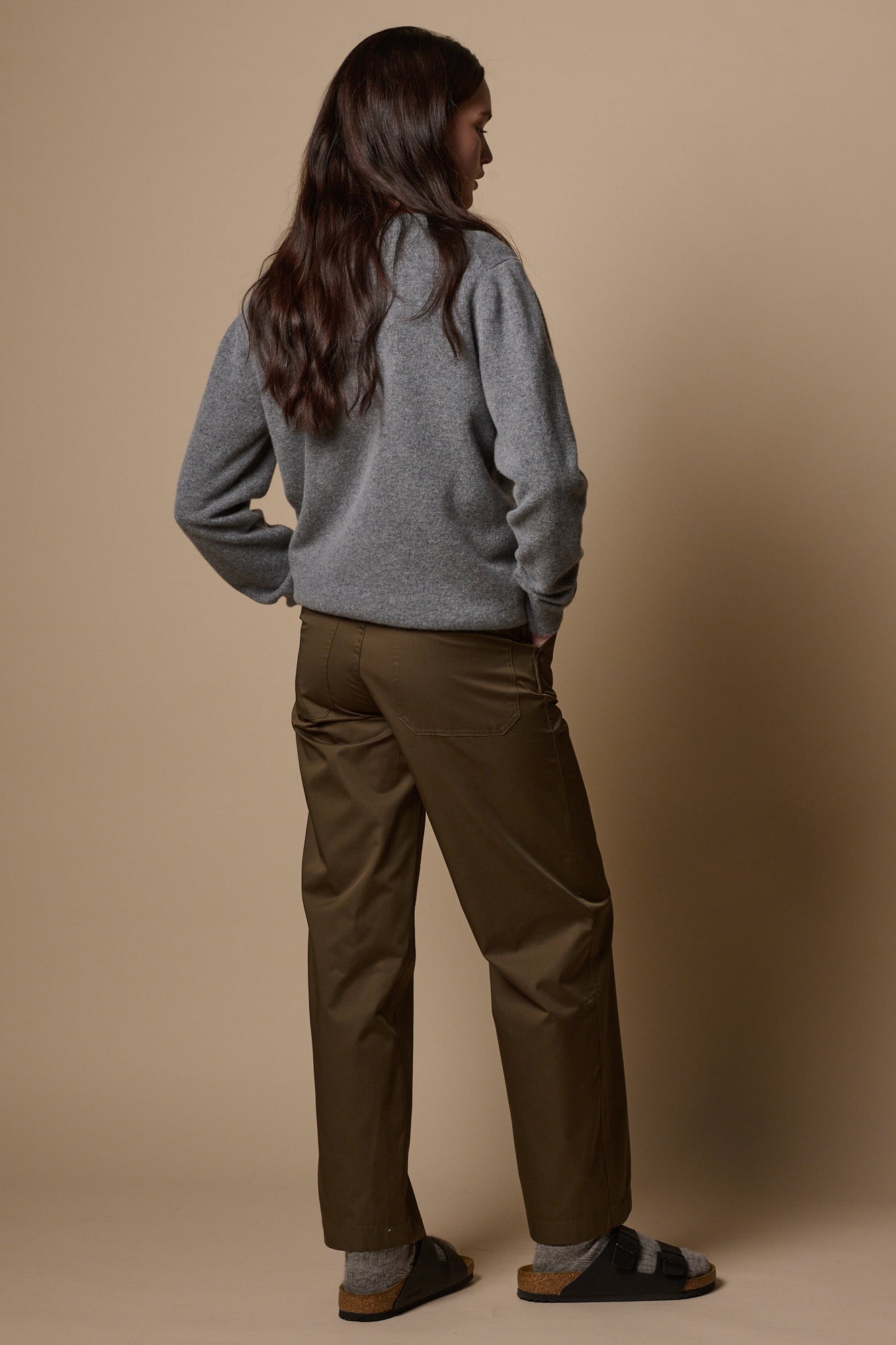 Full body shot of the side of brunette female wearing work trousers in olive with hand in front pocket. Worn with lambswool crew neck jumper