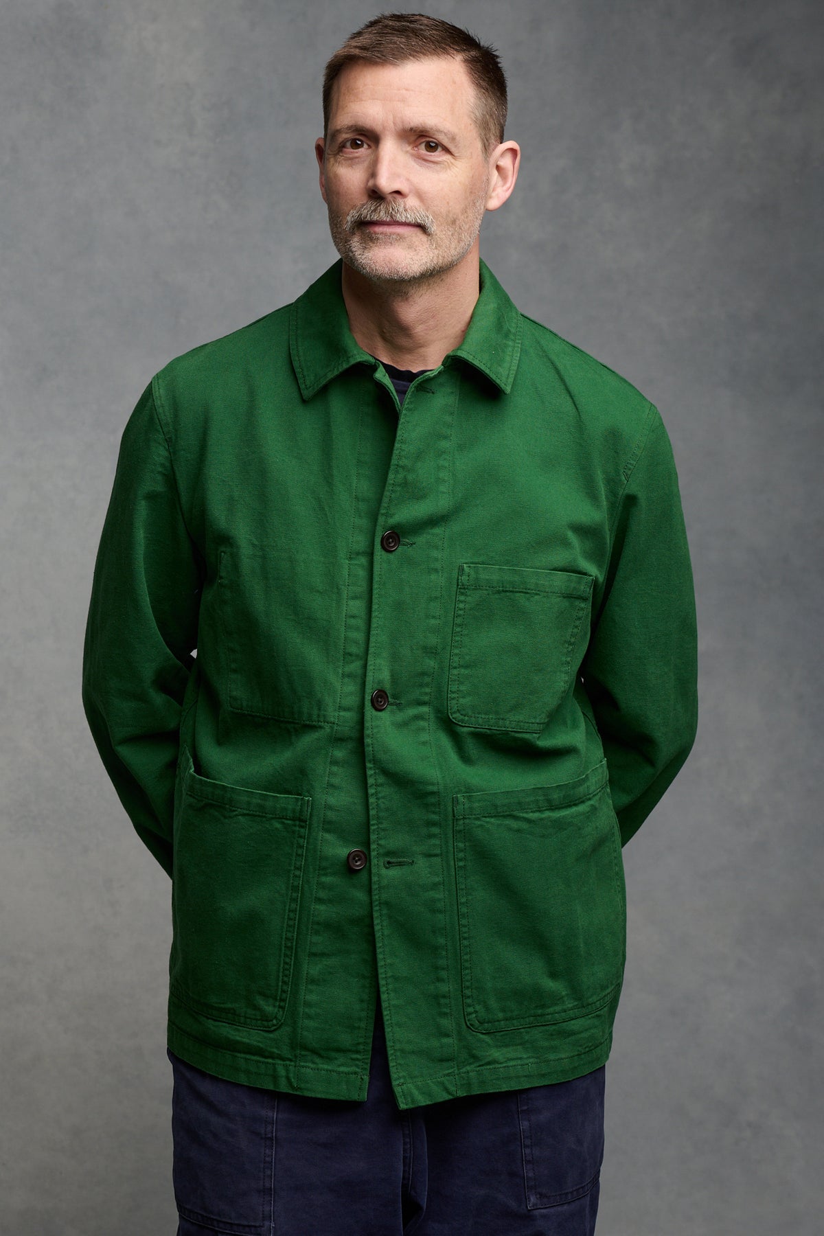 The front of our founder Patrick Grant wearing men's chore jacket in bottle green with two front buttons fastened, his hands held behind his back. Chore jacket worn with cameraman pants in navy