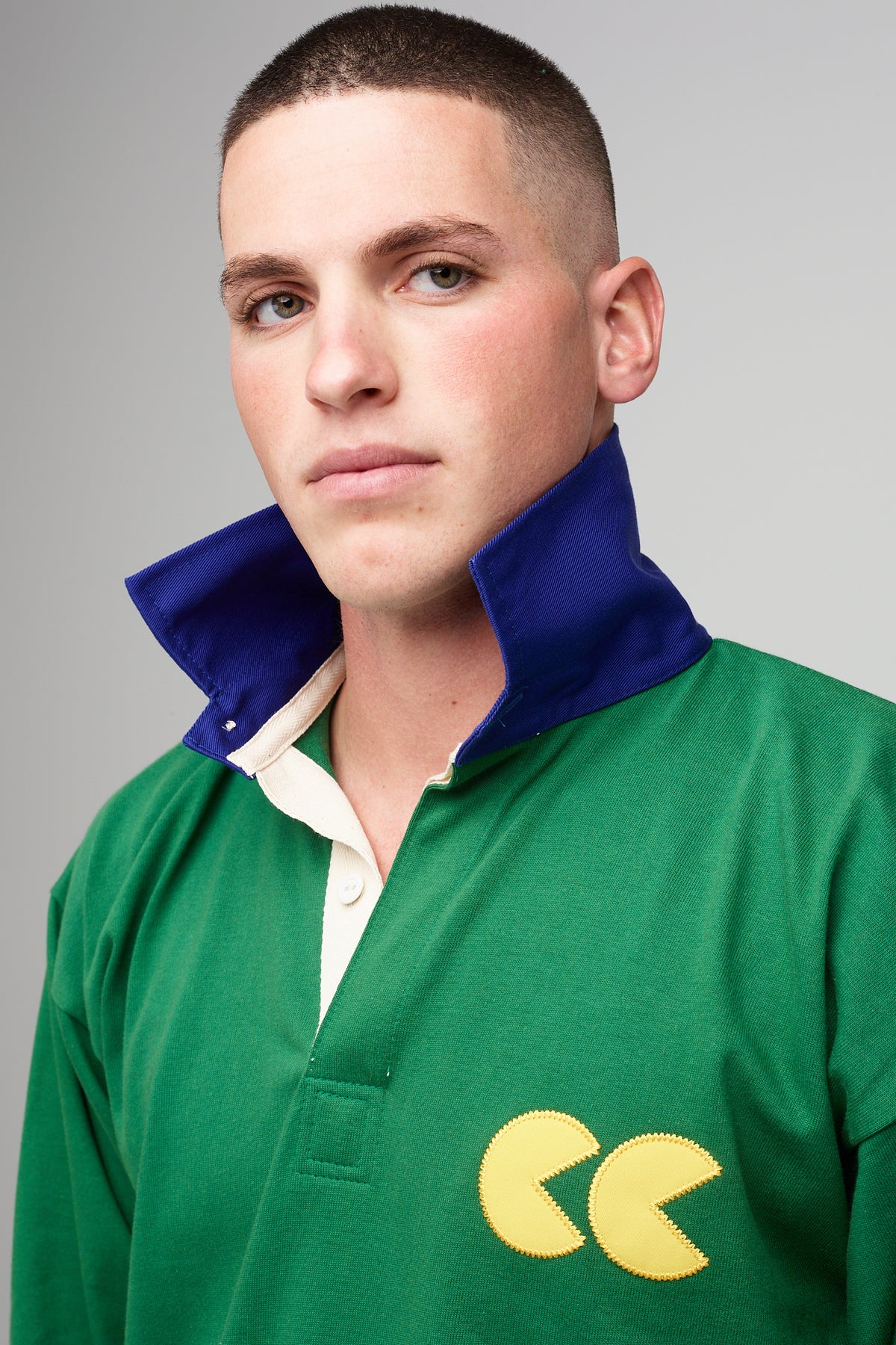 White, brunette male, wearing green rugby shirt with contrast blue collar turned up, yellow CC logo badge