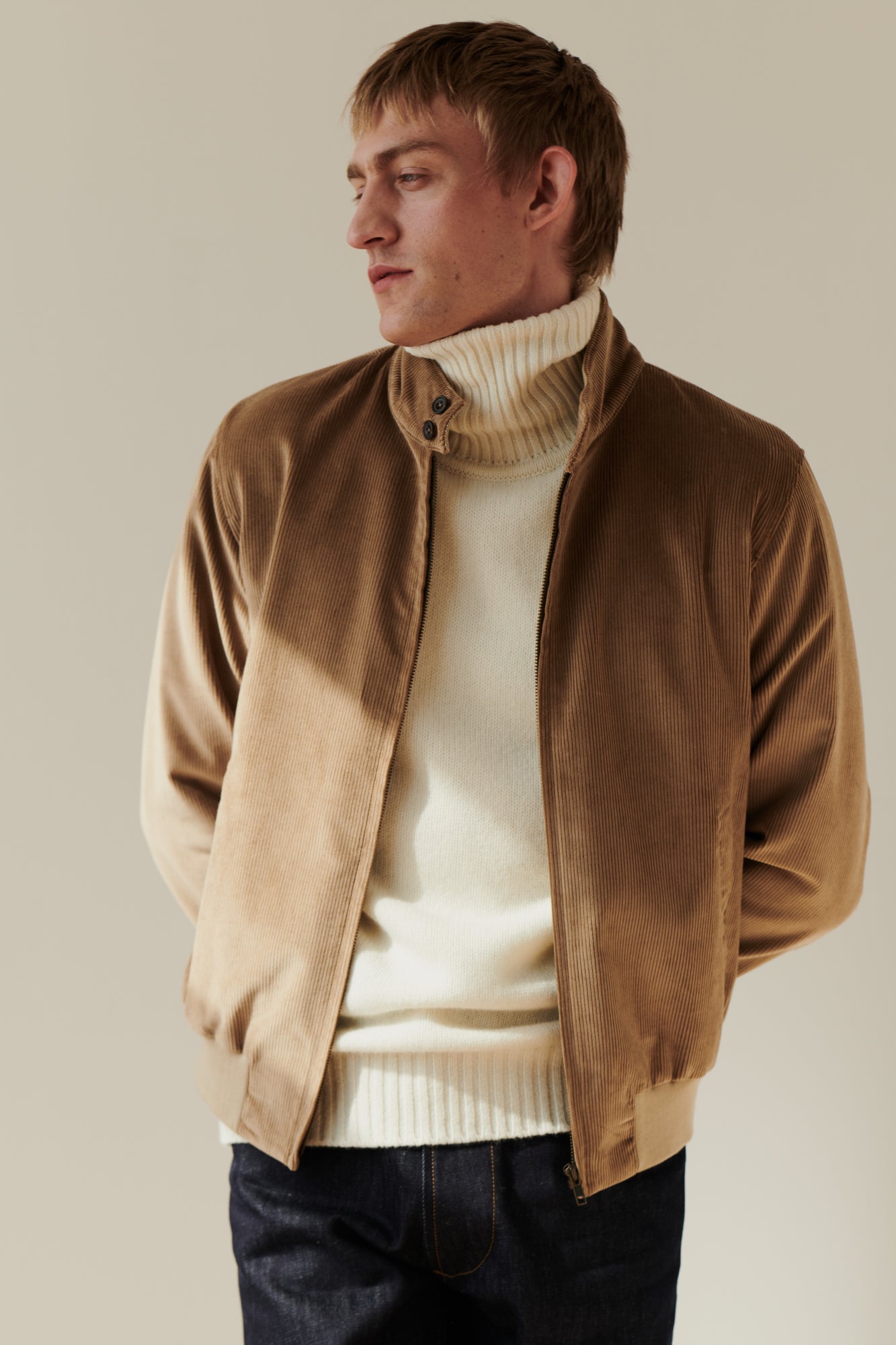 Blonde, white male wearing men's corduroy harrington jacket in stone layered over lambswool roll neck jumper in ecru