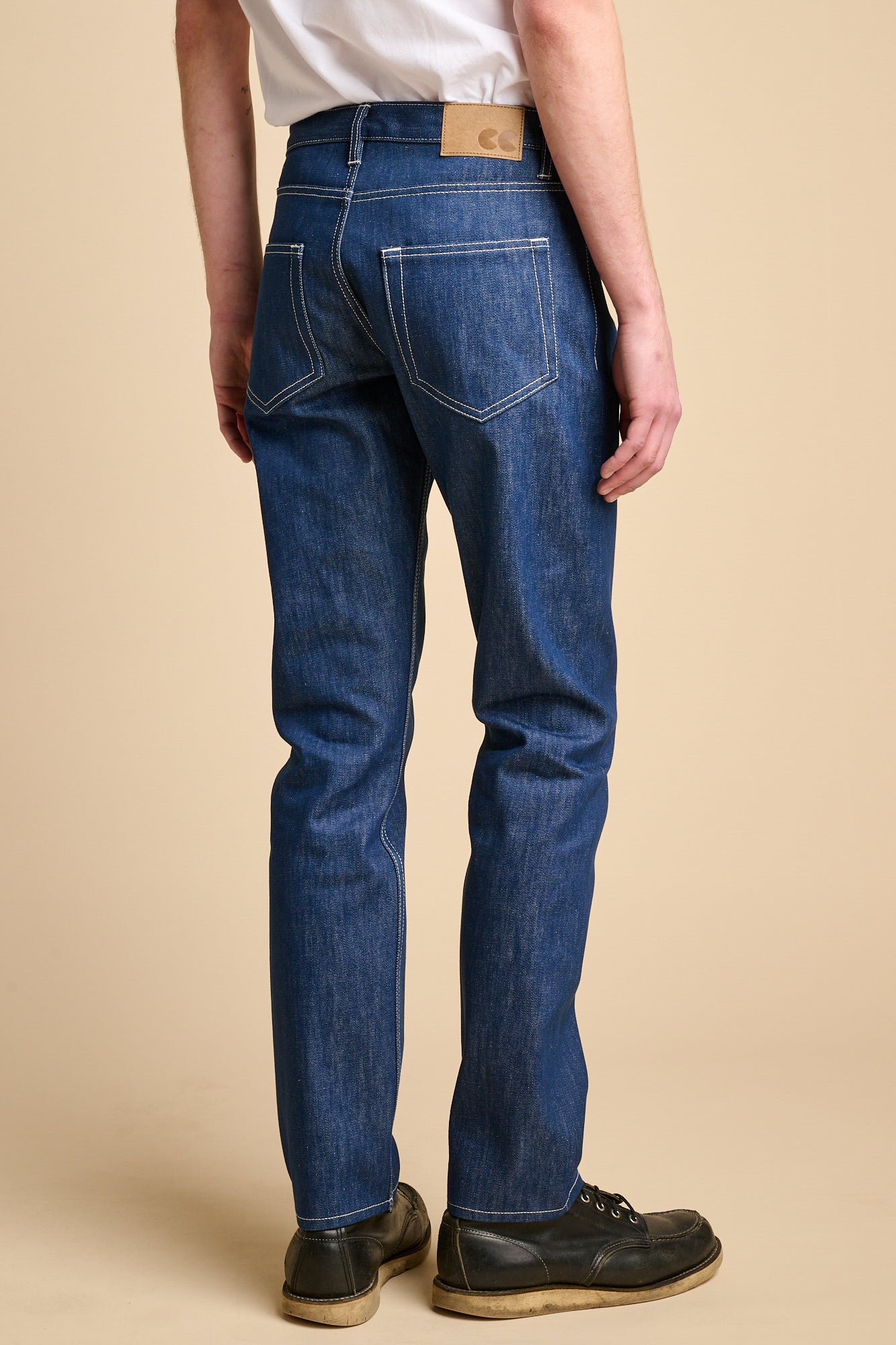 The back of man wearing straight cut jeans in blue with contrast ecru stitching and CC logo jeans patch.