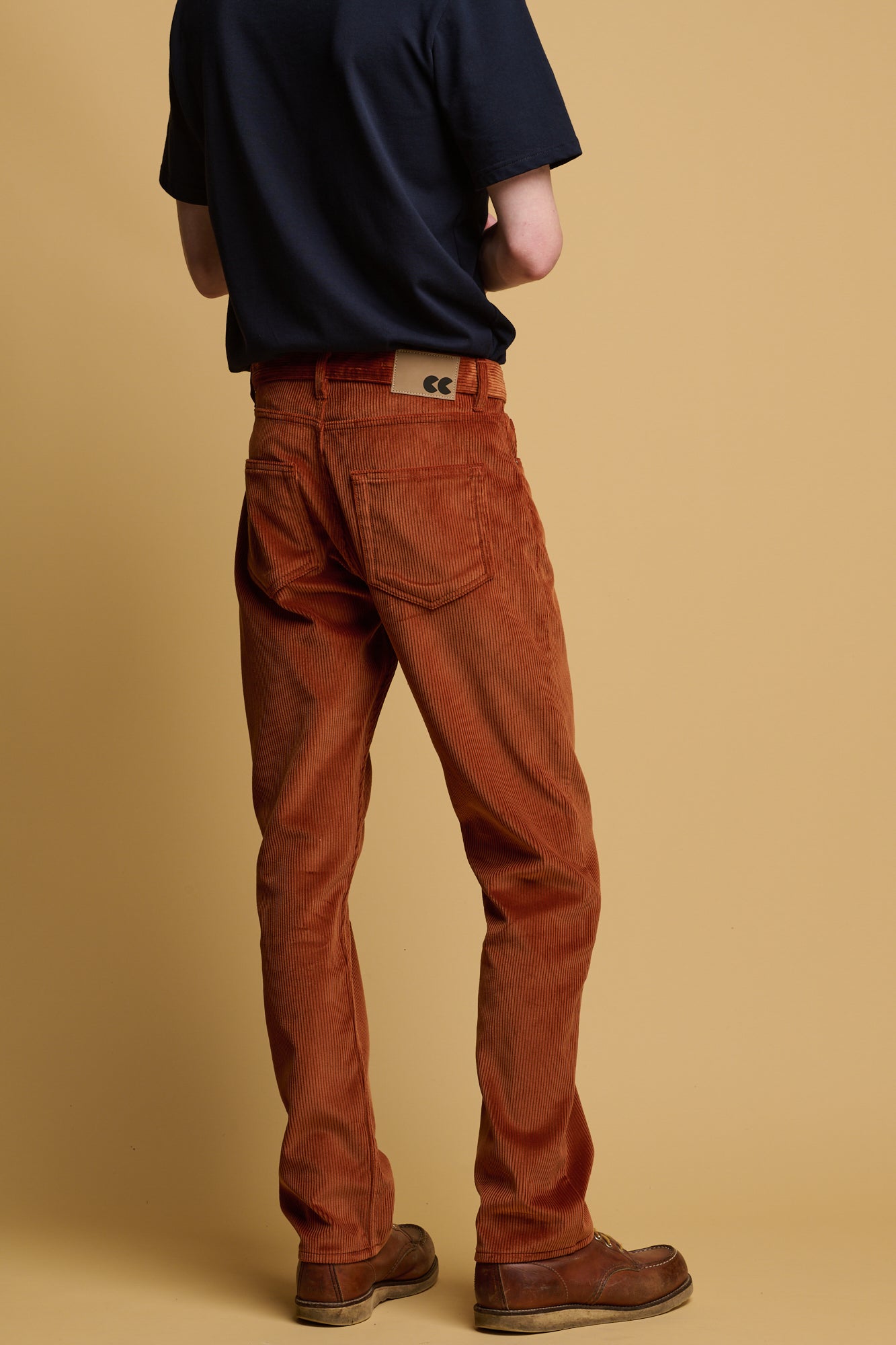 The back of male shoulder down wearing five pocket cord trousers in cinnamon paired with navy t shirt. two back pockets and CC logo jeans patch on waistband with belt loops
