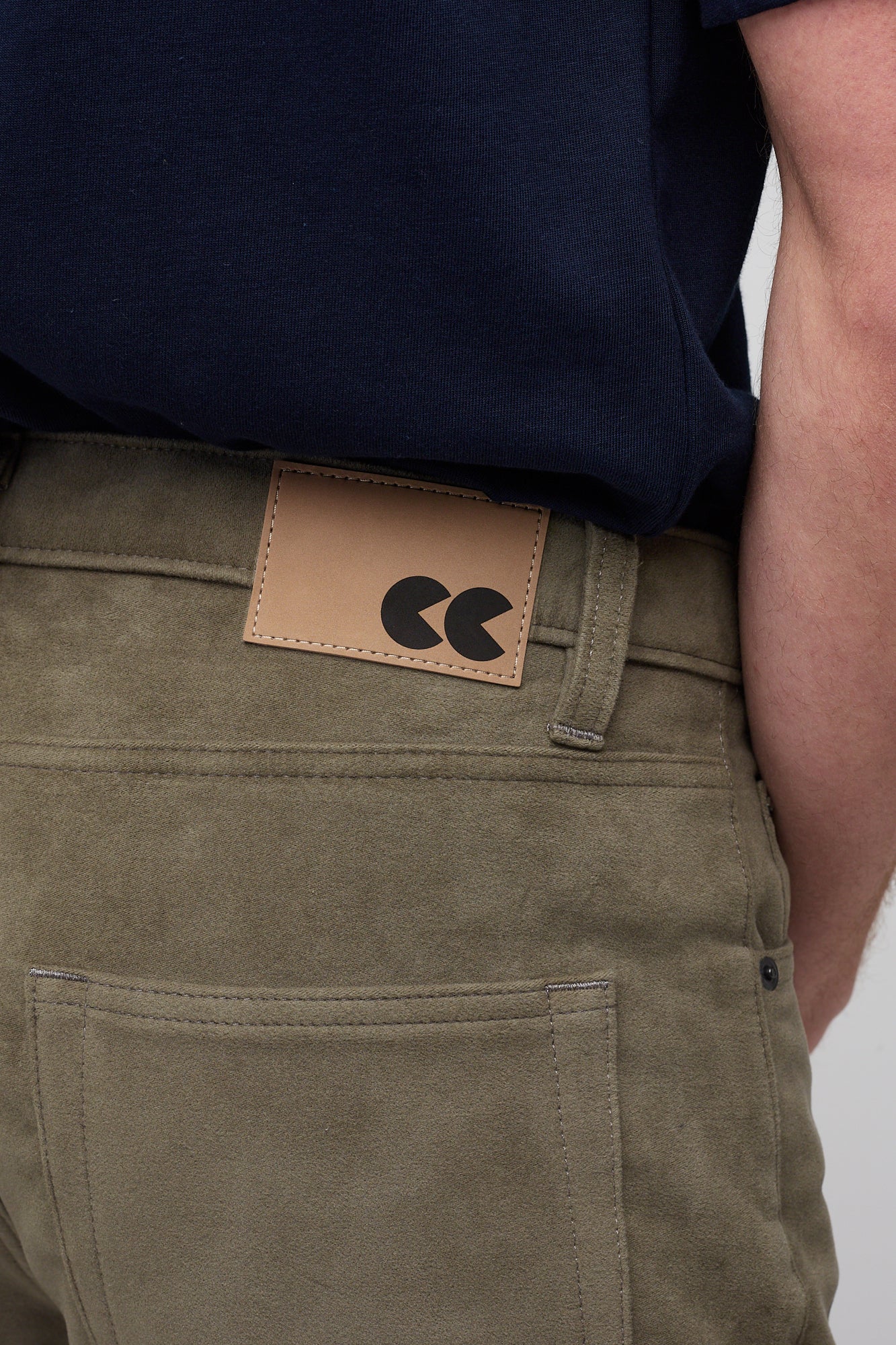 Brunette male in Olive Five Pocket Moleskin Jean styled with navy men's short sleeve t-shirt, brown CC logo badge close up