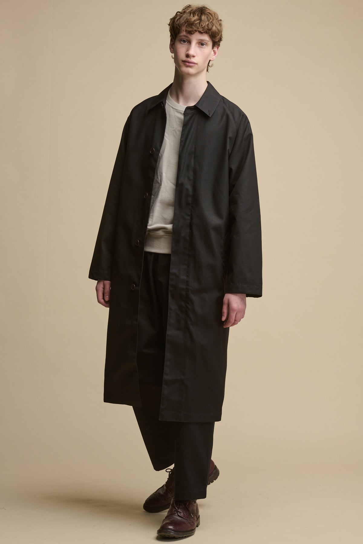 White male with orban hair wearing men's Frank Belted Raglan Raincoat in black, unbuttoned worn over stone raglan sweatshirt and black cameraman pants