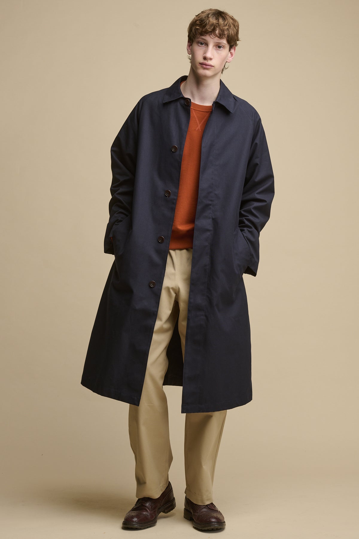 Full body image of the front of male wearing unfastened Frank raglan belted Raincoat in navy worn over raglan sweatshirt in cinnamon and cameraman pants in putty 